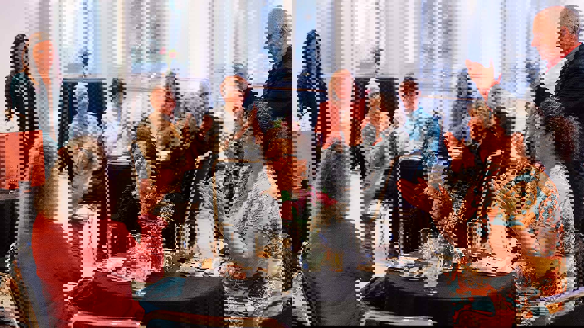 A group of people sitting a smaller tables clapping. There are plates of food and flowers on the tables.