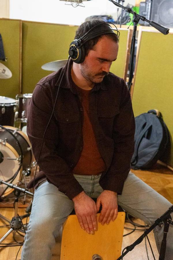 A man with light complexion and short brown hair seated on a cajon and playing with his hands.