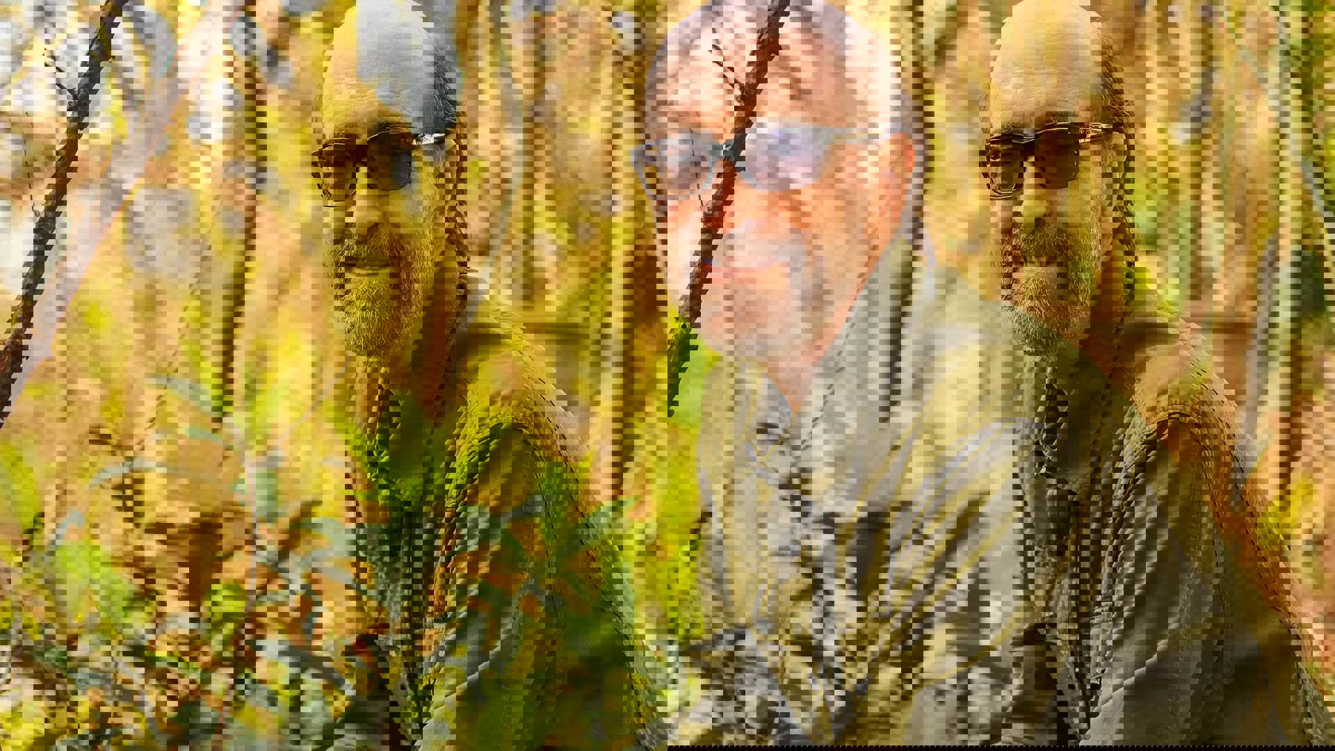 George Winston smiling towards the camera in an outdoor setting. He is a caucasian man with a bald head, grey beard, and glasses