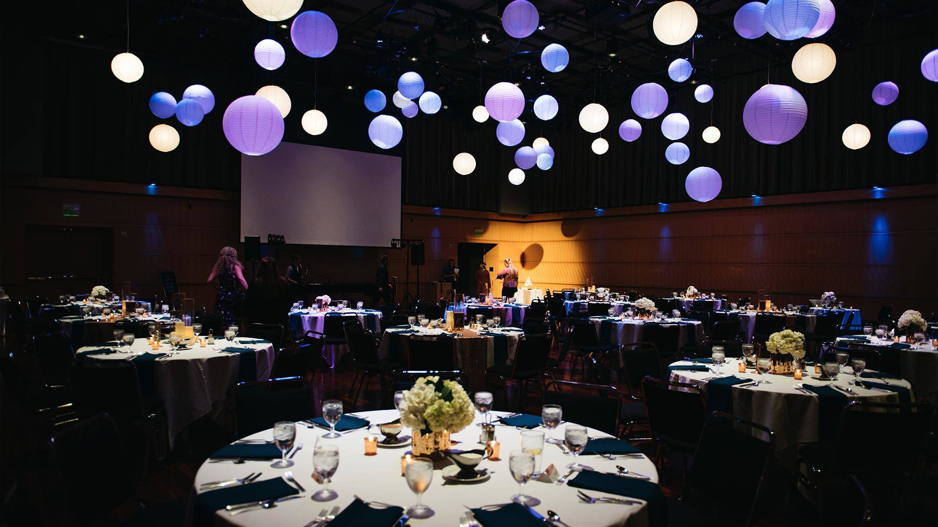 A wedding reception room with tables and spherical purple lamps hanging from the ceiling.