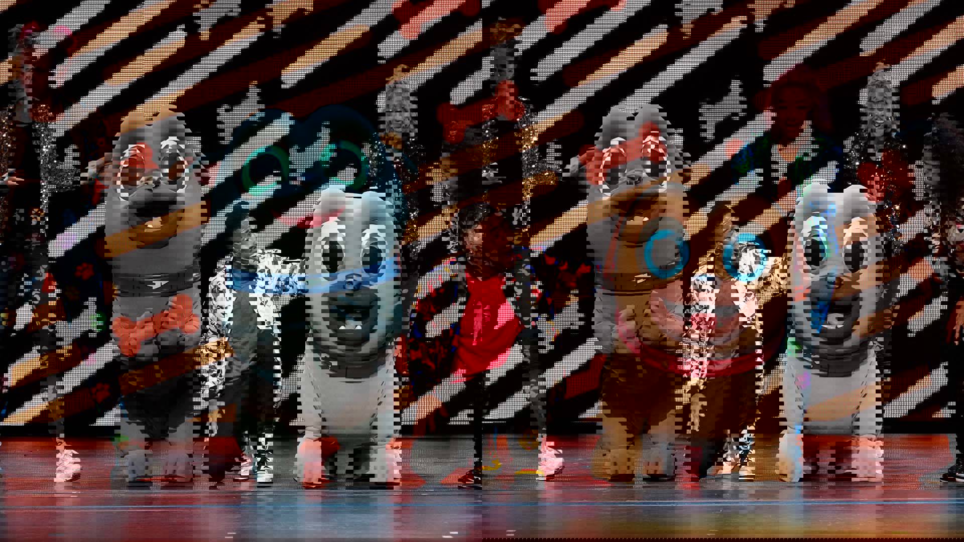 Two large dog costumes with smiling faces next to a young African American man kneeling and talking to them. He has a jacket and red tshirt on.