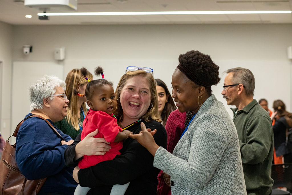 A group of people smiling and talking to each other. One woman is holding young black child in a red shirt.