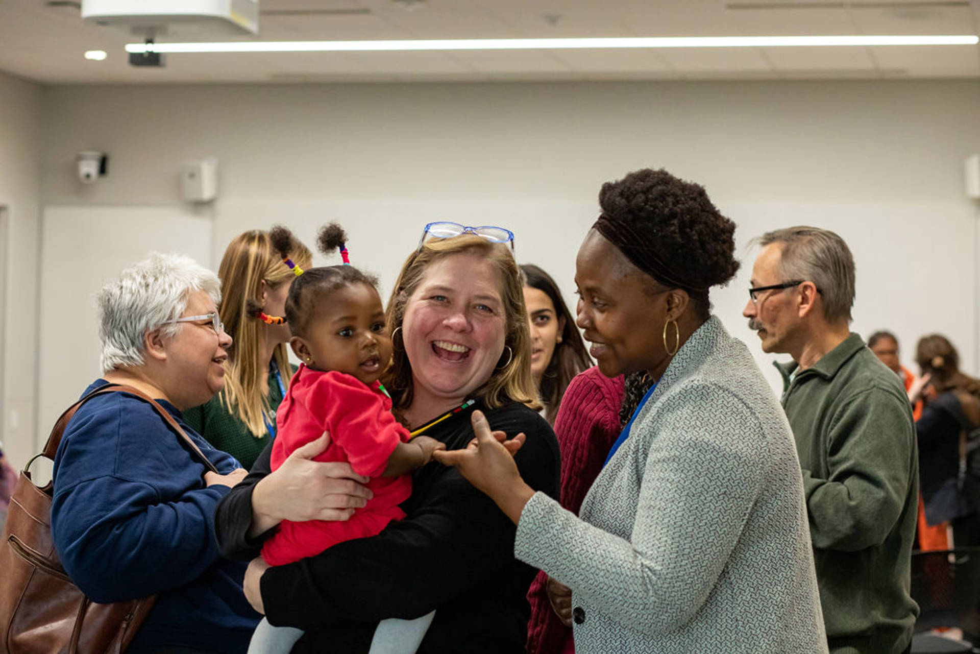 A group of people smiling and talking to each other. One woman is holding young black child in a red shirt.
