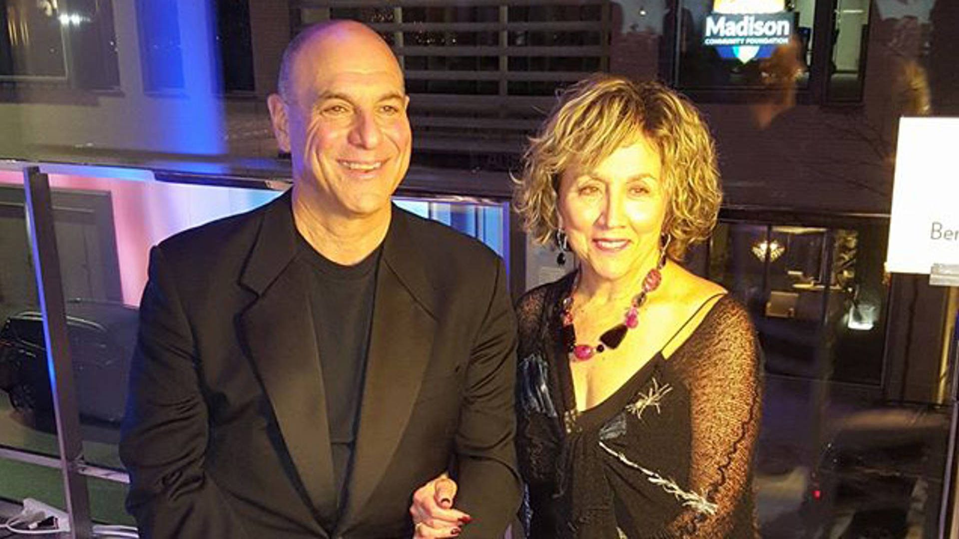 A bald middleaged caucasian man in a tuxedo jacket and black t-shirt posing for a photo with a caucasian middleaged woman with medium dirty blond hair in a black dress.