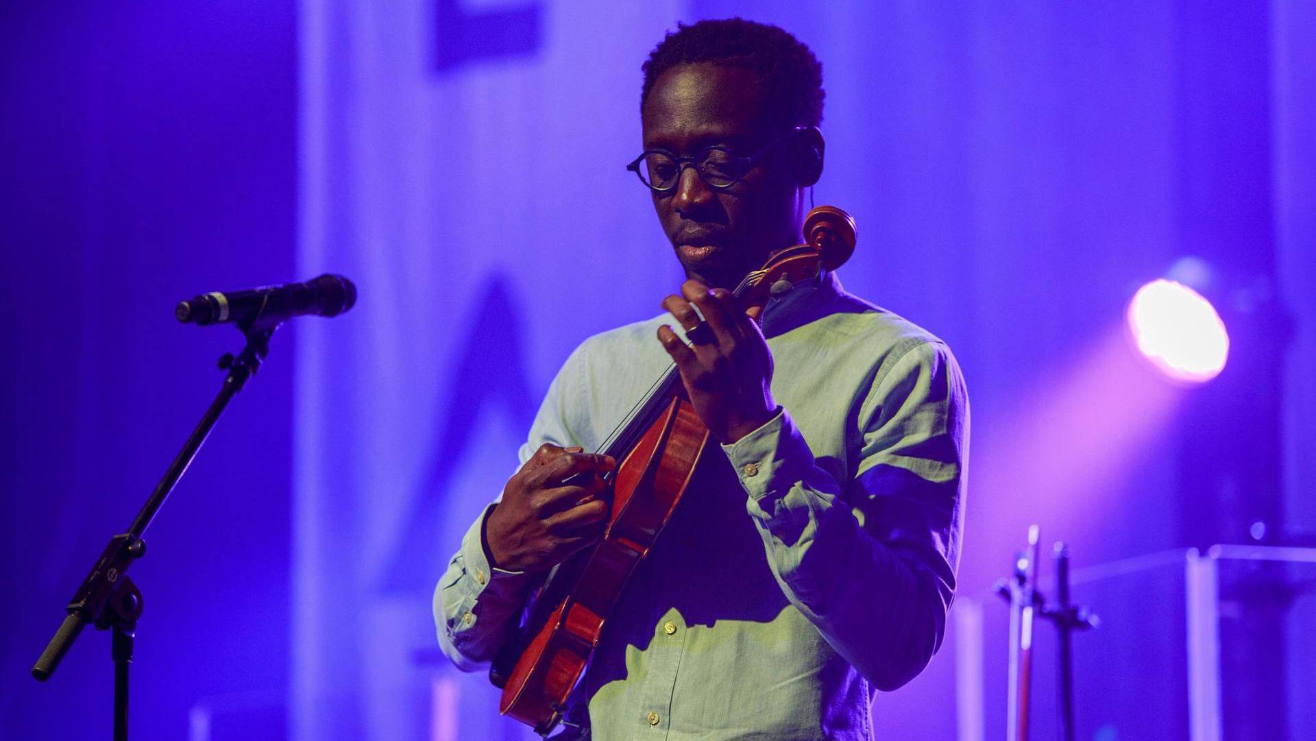 A man with dark complexion, a light shirt, and short hair plays a violin, holding it like a guitar.