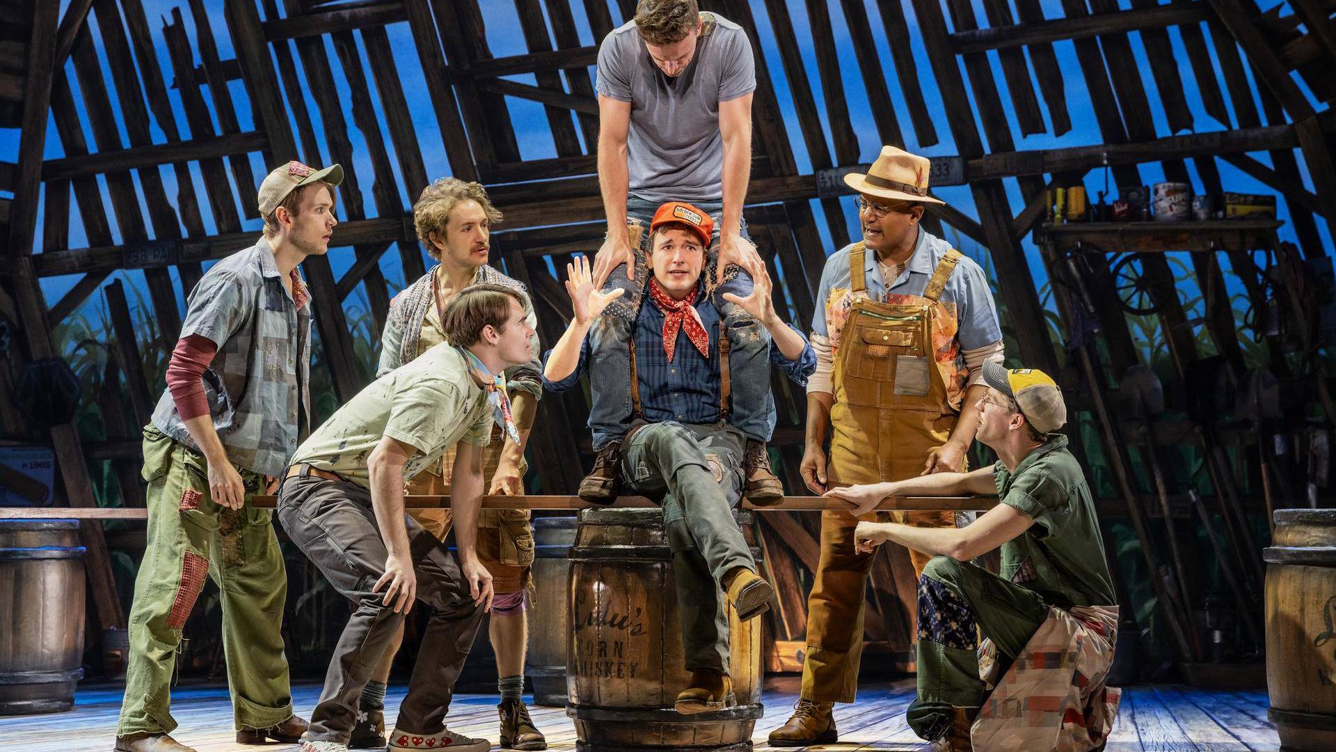 A group of men in working farm outfits huddle around a man with an orange cap, neck bandana, and blue jacket holding his hands up as he sings. Another man is sitting on his shoulders.