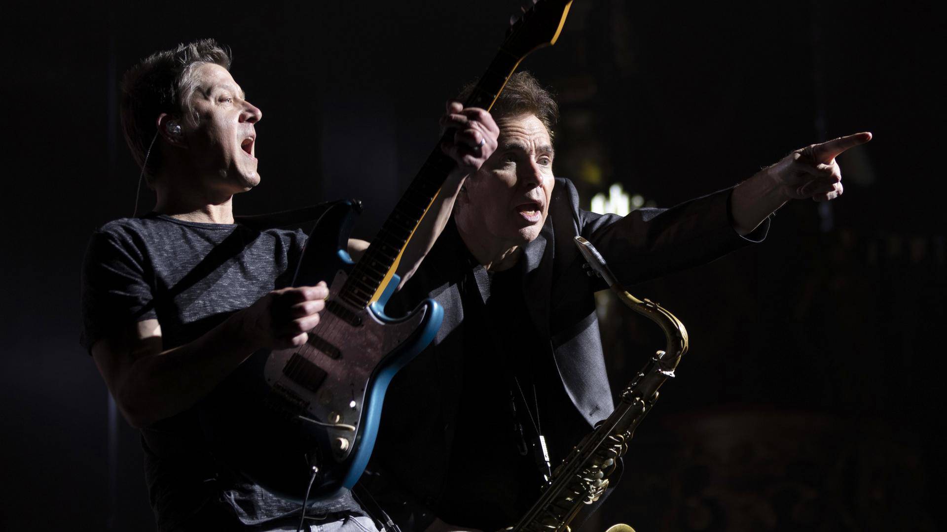 Two white men. The man on the left is holding a blue guitar and yelling. The man on the right is holding a saxophone and pointing.