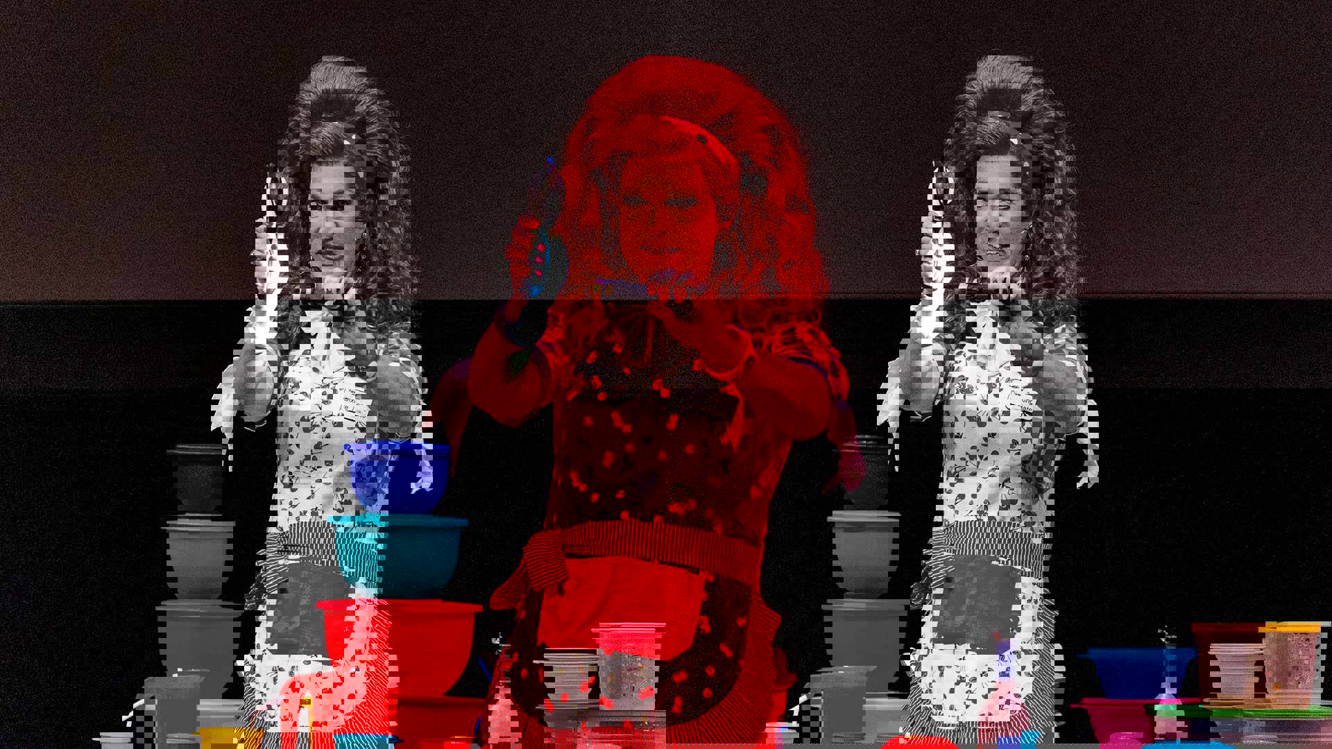 A drag performer with light complexion, voluminous red hair, red lipstick and large diamond drop earrings performing on stage with colorful Tupperware bowls, wearing a red and white gingham dress and cherry print apron.