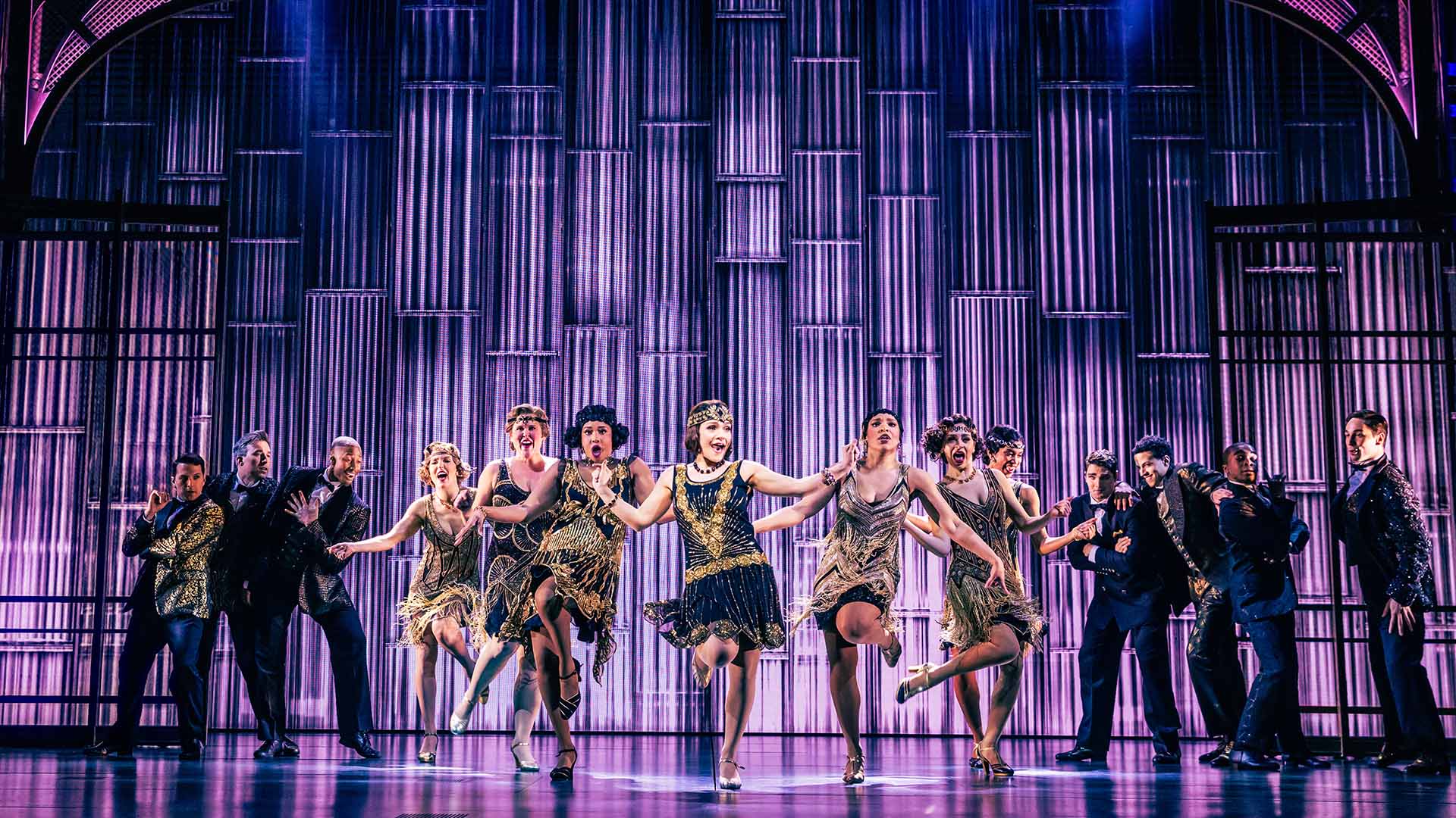 Seven women in flapper costumes dance against a purple art deco backdrop as men wearing suits stand on either side of them watching.