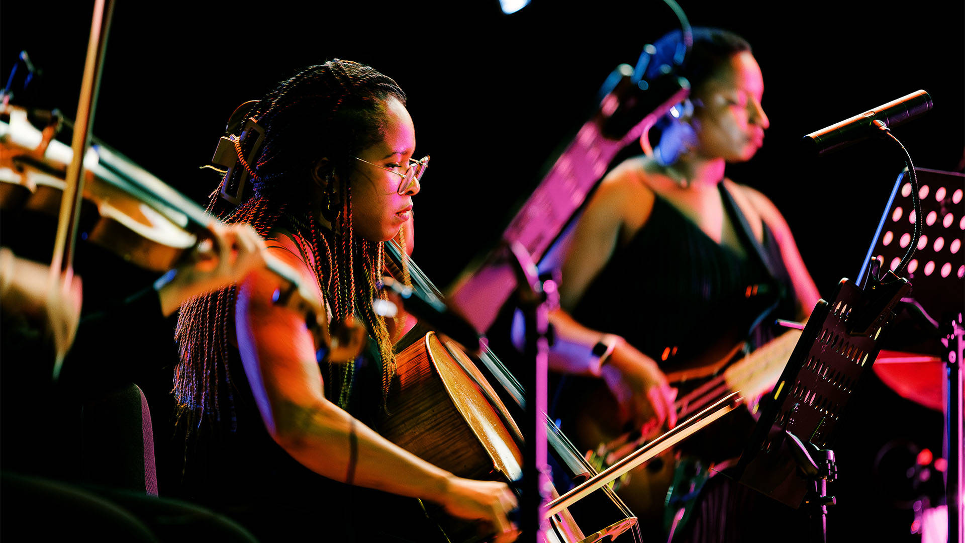 A woman with dark complexion and dark hair with a dark top playing a cello.
