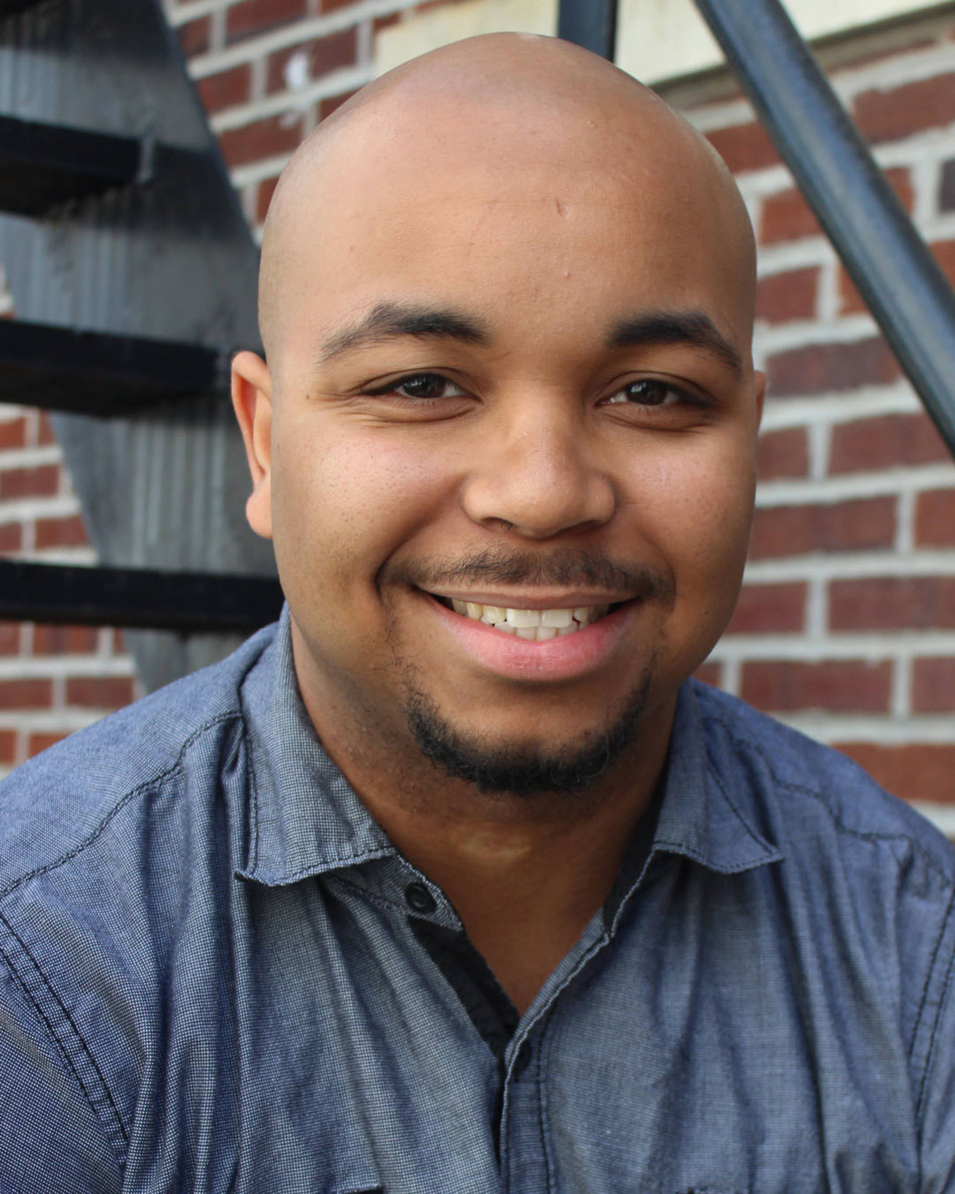 Headshot of a man with dark complexion, a bald head, brown eyes and a goatee.
