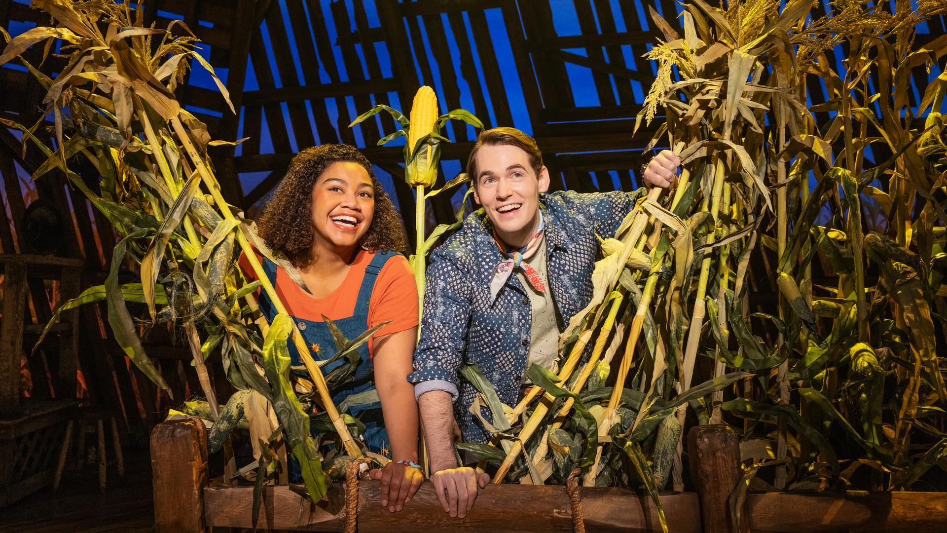 Two people are standing and smiling surrounded by a cornfield with one hand on a wooden fence. They are holding a stalk of corn together in their hands.