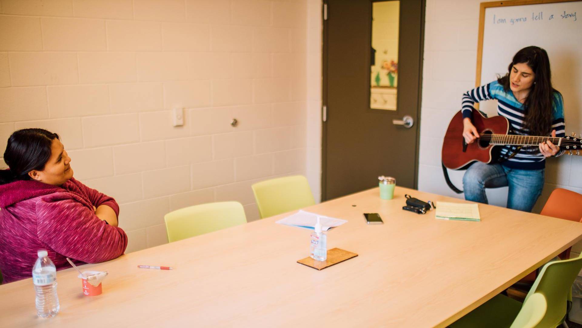 A woman with medium complexion and long brown hair playing guitar and singing at the head of a long beige table. Another woman with medium complexion and brown hair is sitting listening.