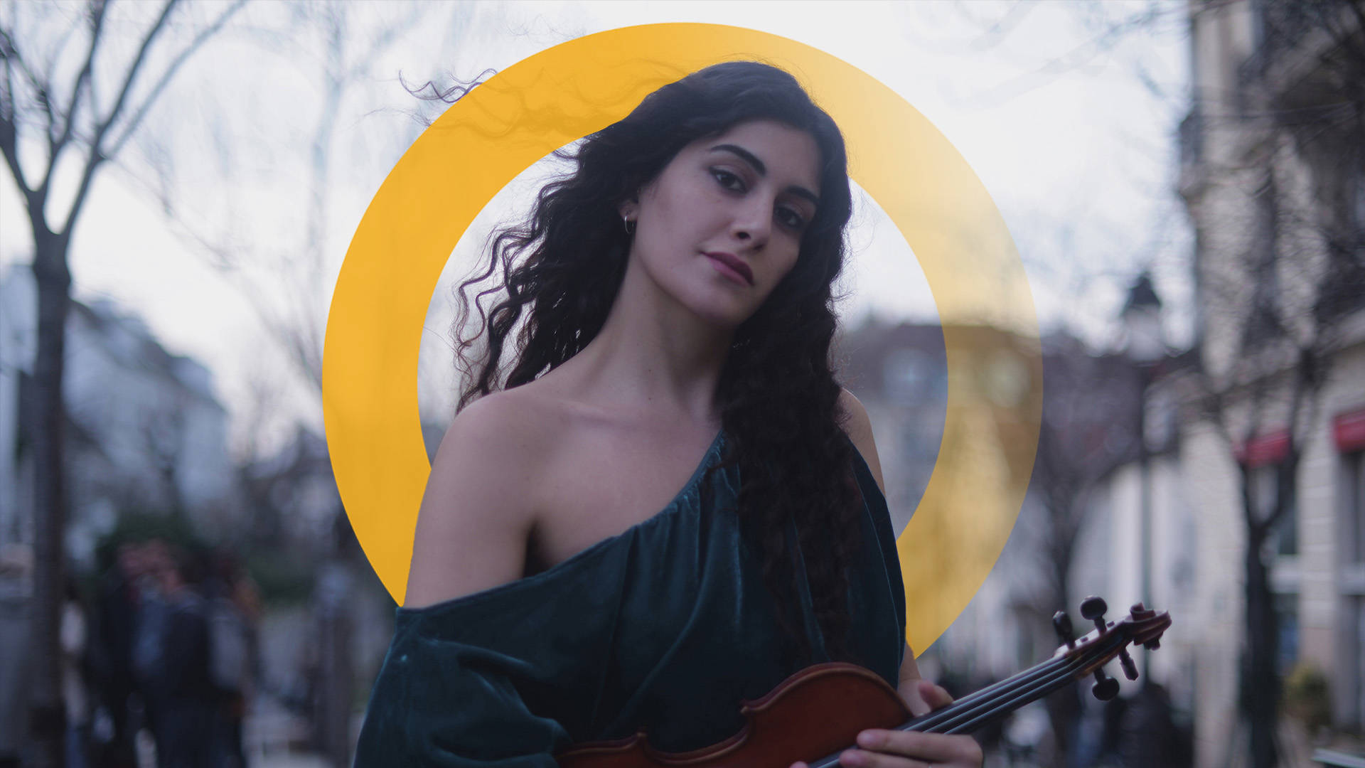 A woman with long, wavy, dark hair wearing a green velvet one-shoulder dress. She is holding a violin outside. A yellow "O" is digitally placed around her head.