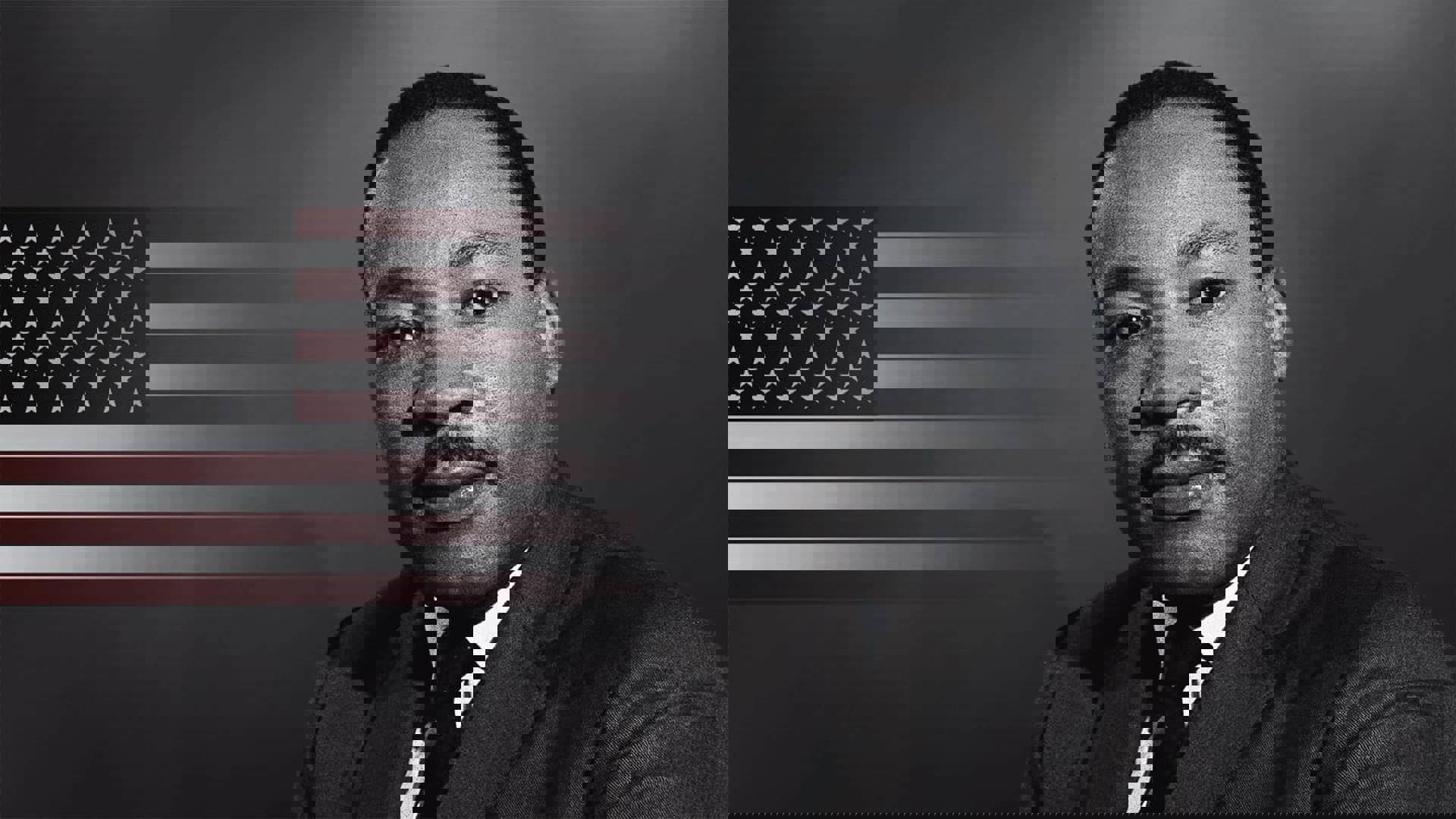 A black and white image of Martin Luther King Jr. A black man with short hair and a mustache wearing a suit. The American flag in color is behind him.