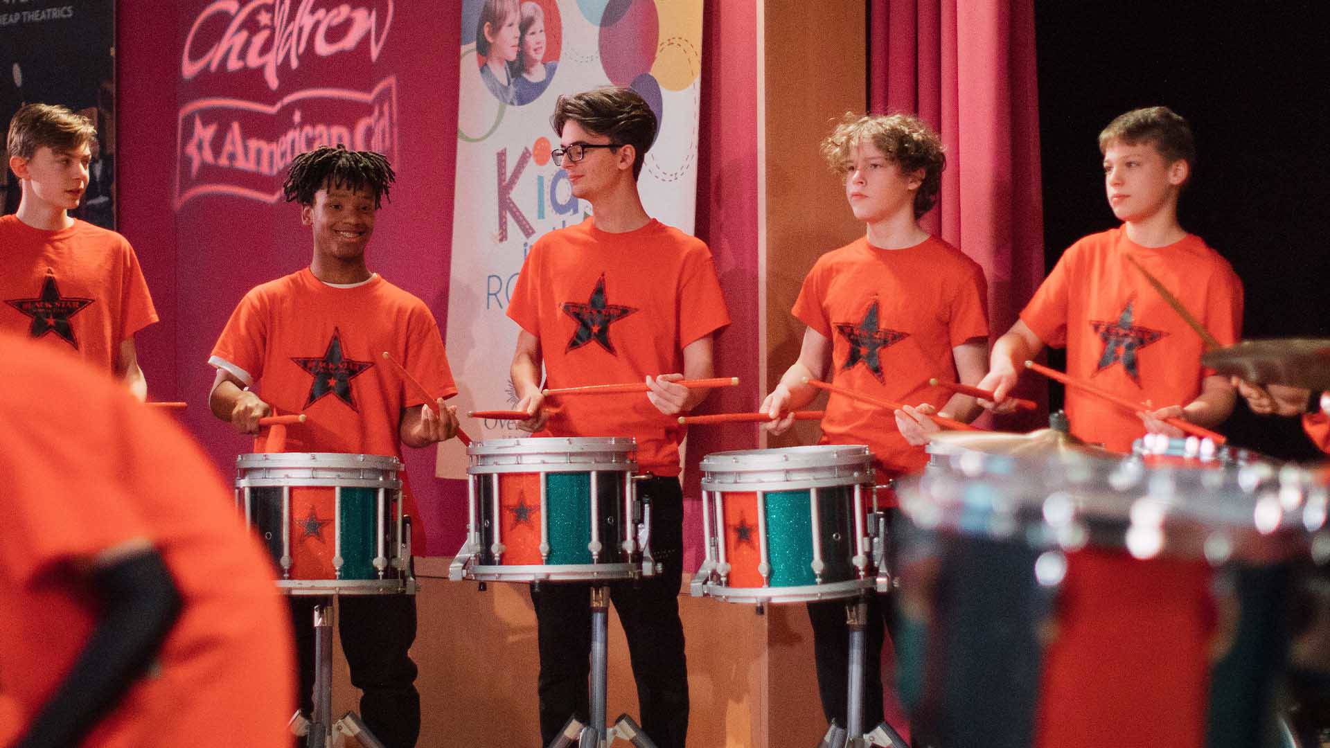 Young adults in orange shirts playing marching snare drums.