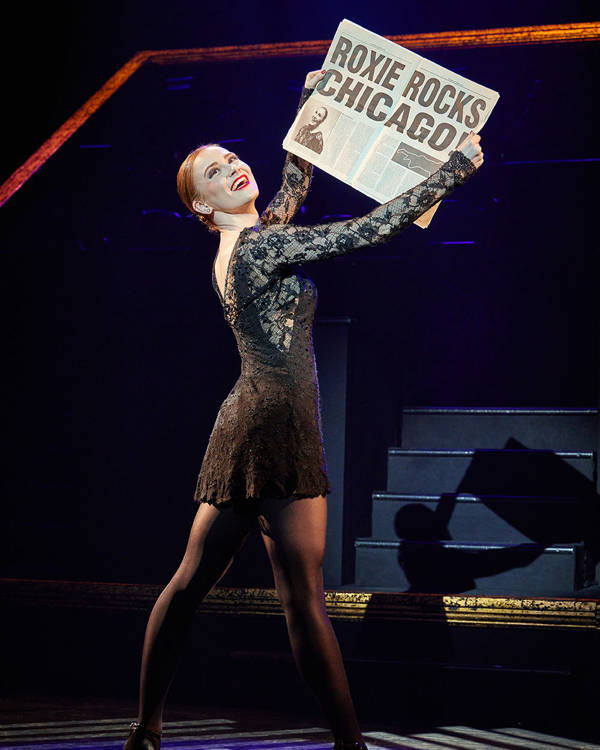 A white woman in a short black dress with her red hair in a tight bun. She is smiling and holding out a newspaper that reads "Roxie Rocks Chicago!"