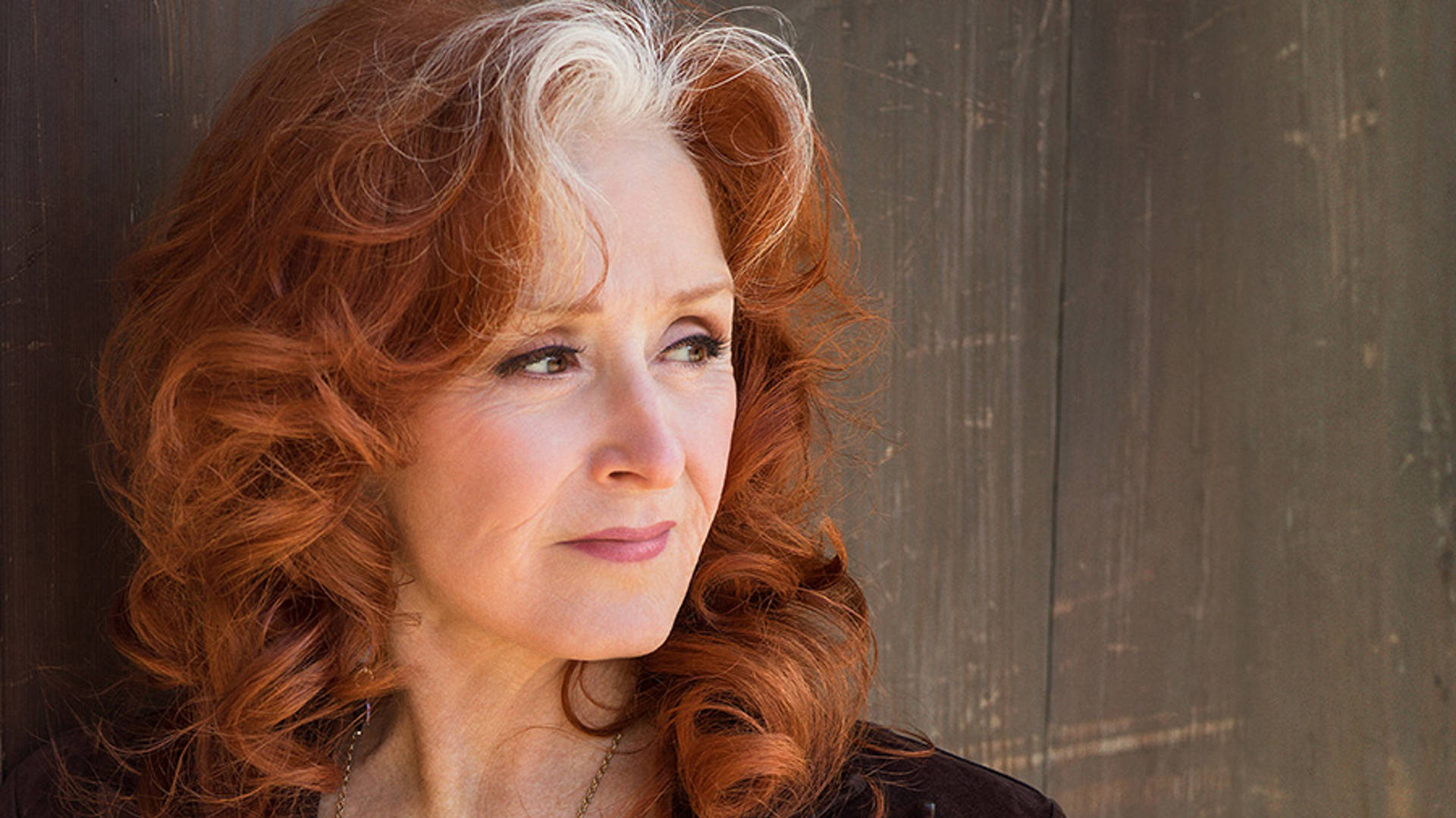 Bonnie Raitt looks to her left. She is standing against a wooden background. She has striking red hair with white highlights and brown eyes.