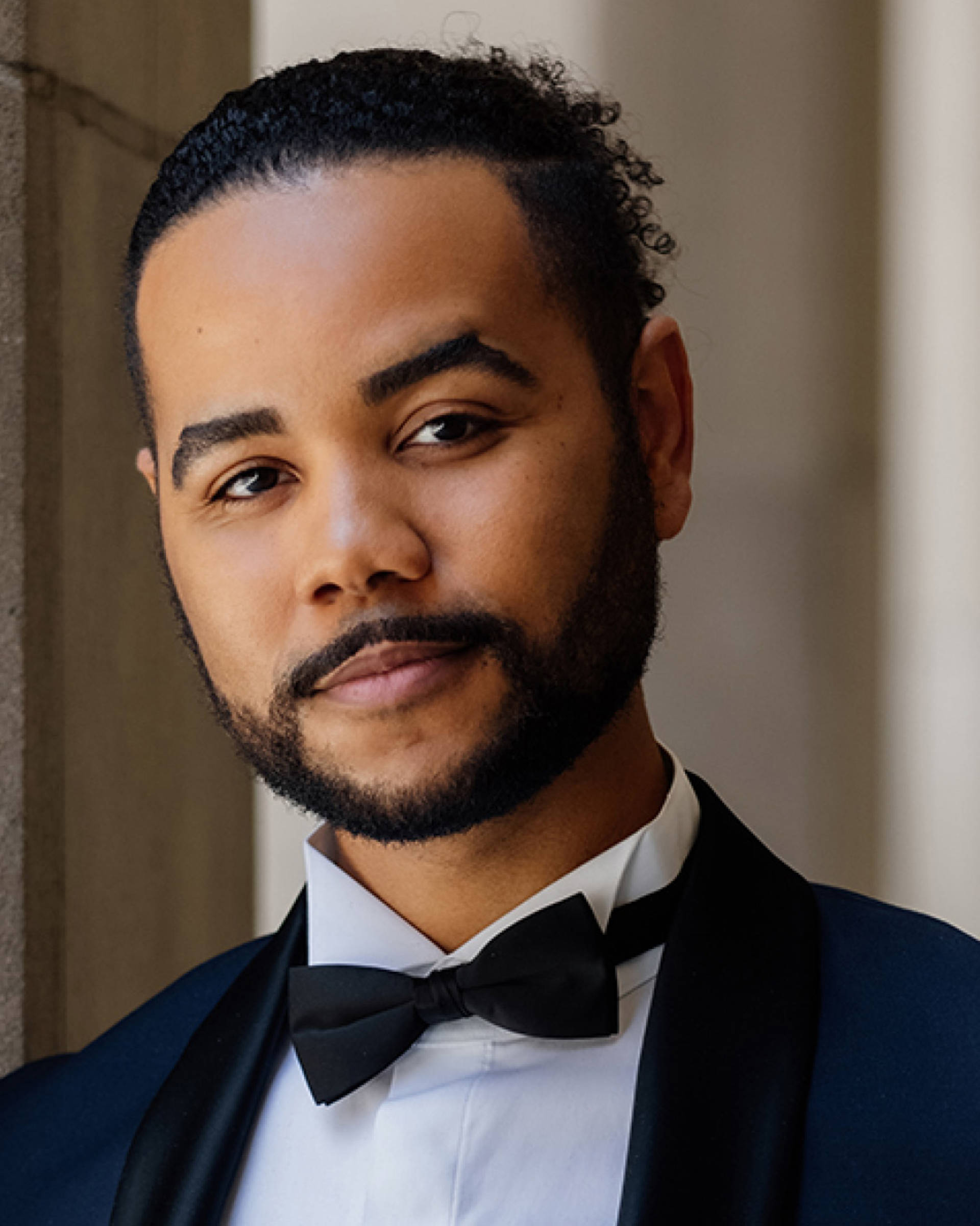 Headshot of a man with dark complexion, dark hair, brown eyes, and a short beard. Hey is wearing a tux.
