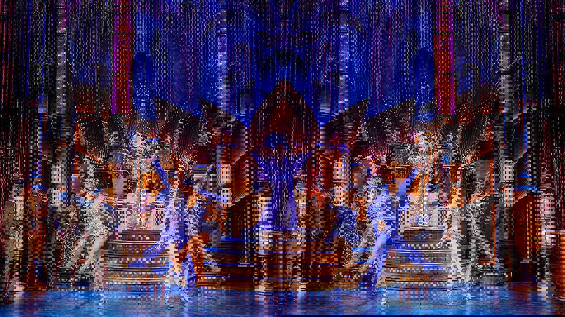 A dazzling stage set-up with art deco decorations as a band plays with a singer at the center wearing a purple 1920s party dress and a large feather headpiece. Other dancers in purple kick and pose off the stage steps.
