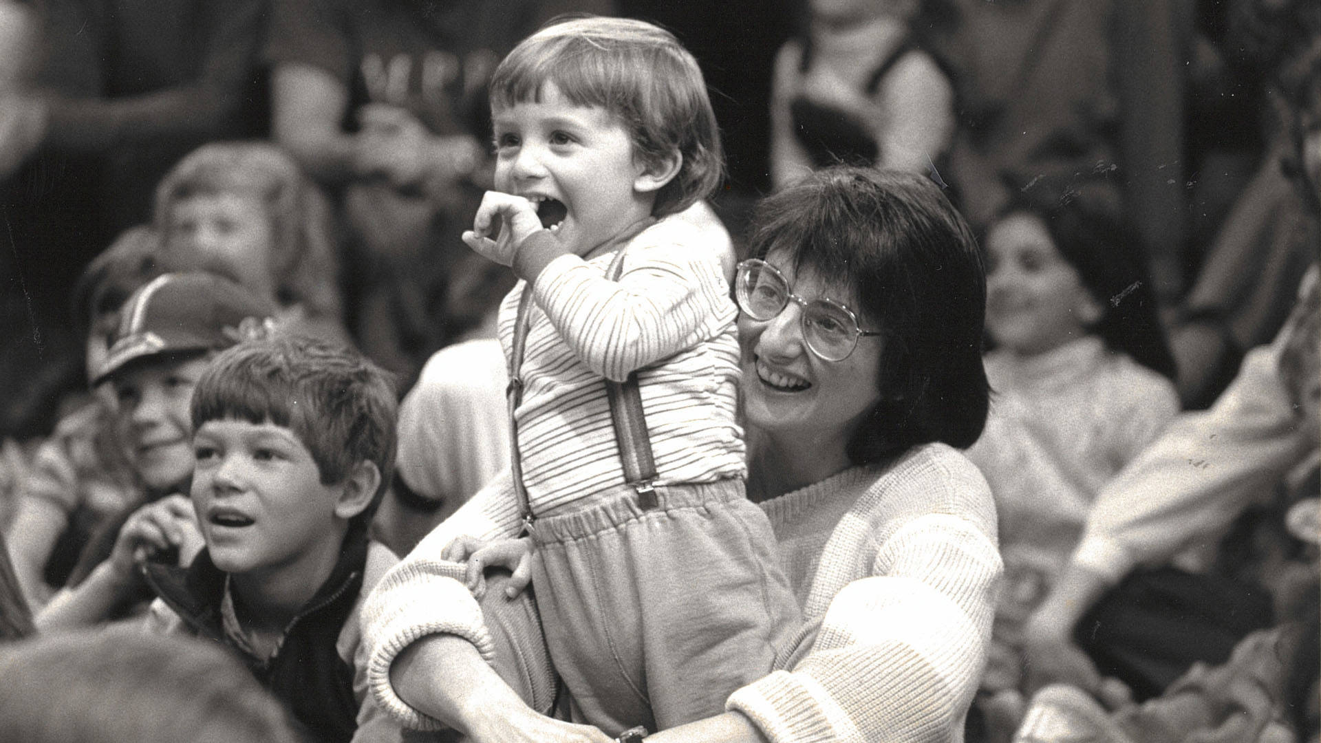 Greyscale photo of a smiling mother with dark medium length hair, light complexion and large glasses. She is a holding a toddler who is also smiling excitedly with a pointer finger in his mouth.