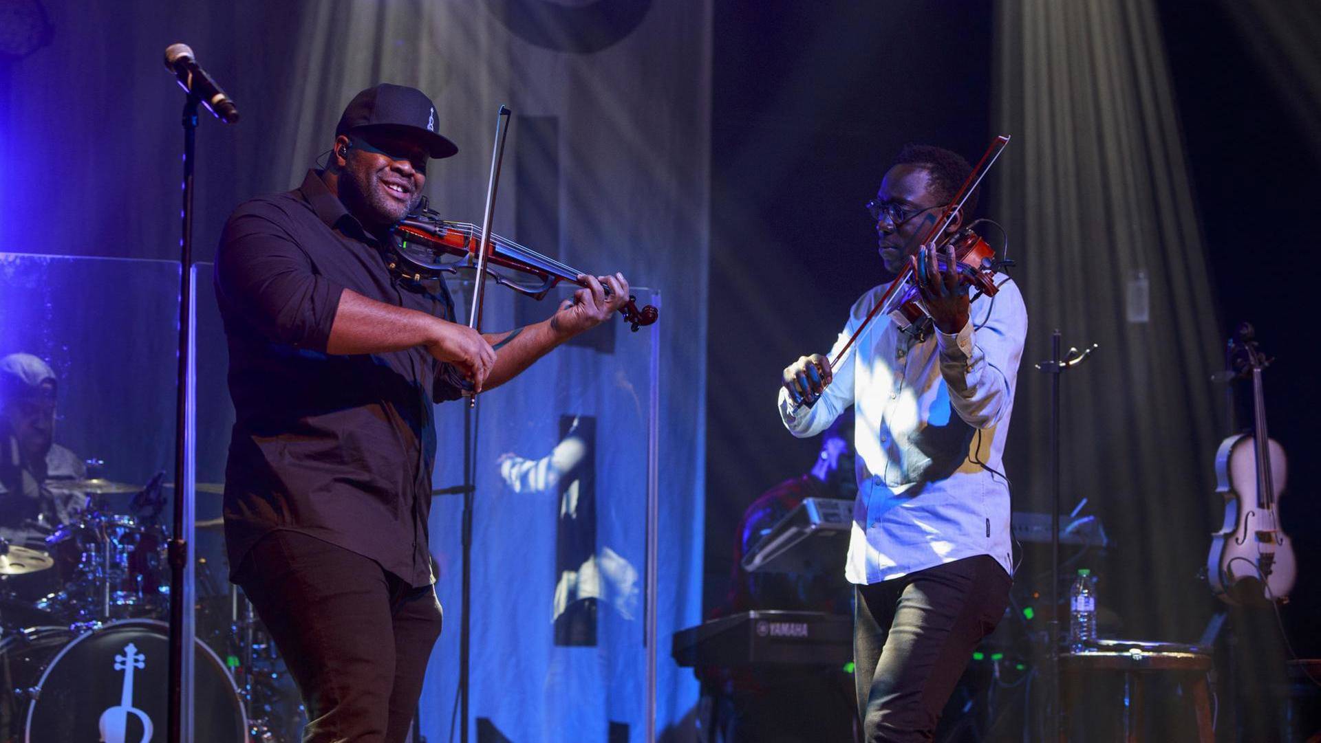 Two men in stylish clothing face eachother and play violins. The one on the left has a black baseball cap, dark complexion and dark clothing. The one on the right has short hair, dark complexion and a white shirt.