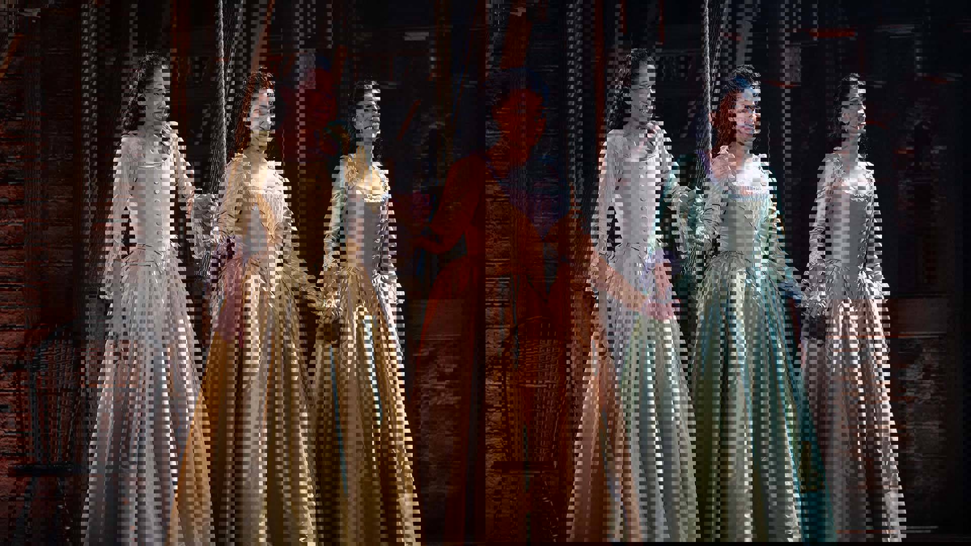 Three women with long hair wearing long gowns hold hands and smile.