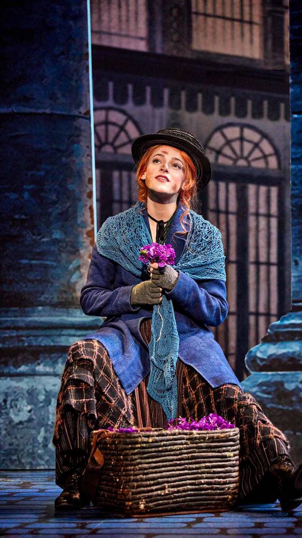 A woman sitting with a basket of purple flowers looks off in the distance. She has red hair, is wearing tattered clothes and has dirt on her face.