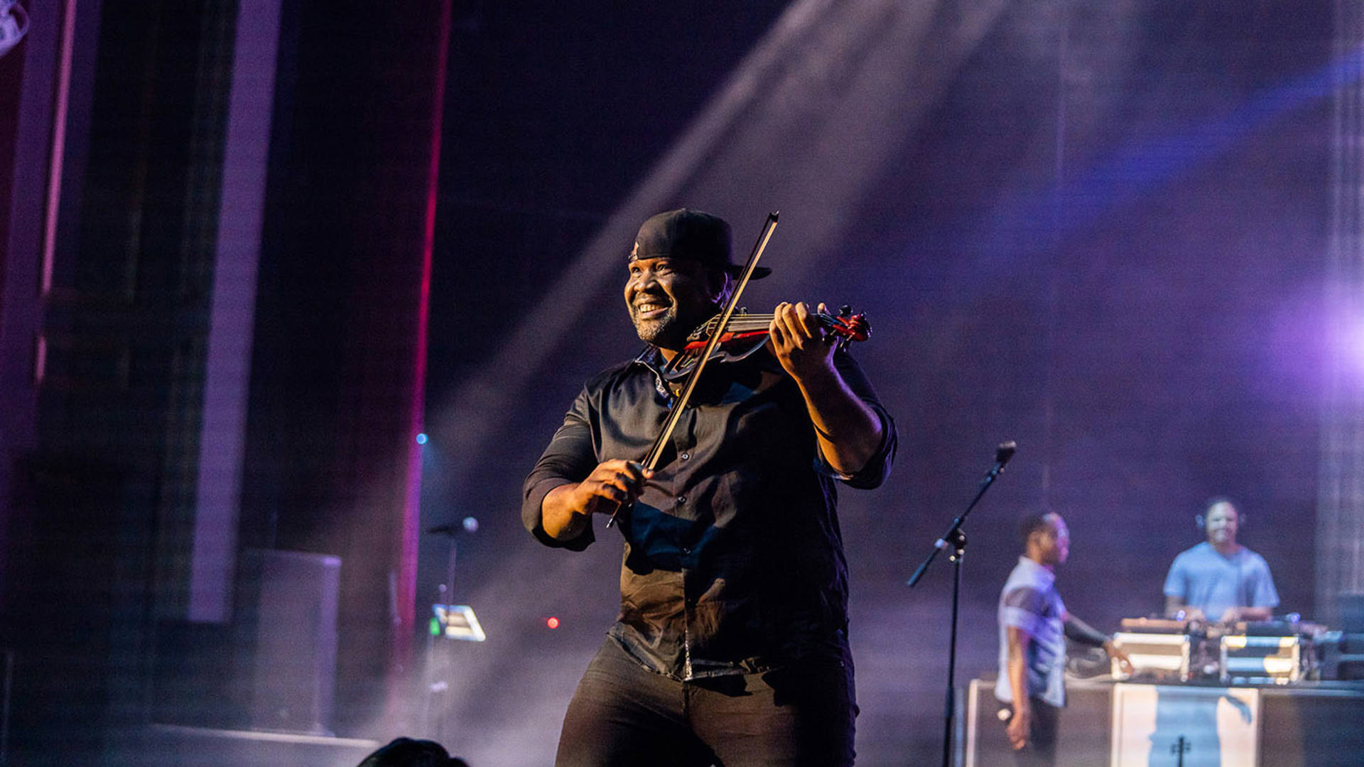 Kev Marcus playing the violin on stage. He is smiling and wearing a hat.