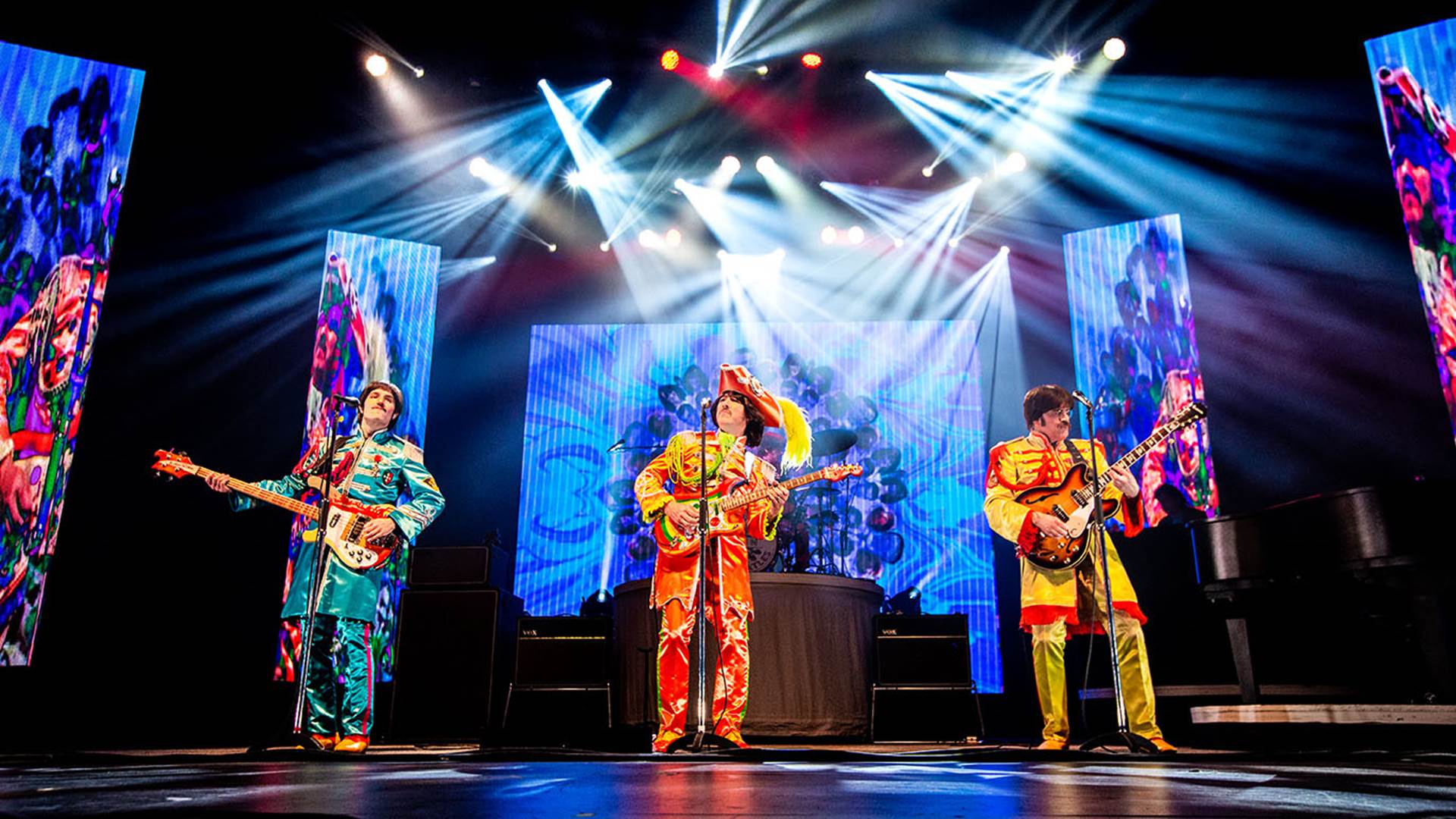 Four Beatles impersonators performing on stage with guitars. They are wearing colorful Seargent Peppers outfits and large projections are behind them.