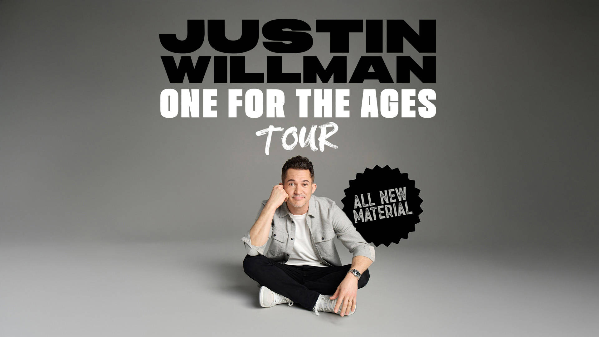 Gray banner that reads: "Justin Willman: One for the Ages Tour. All new material." Sitting below the text is a man with light complexion, short dark hair, wearing a grey shirt and black pants. He is cross-legged with one hand on his cheek, smiling softly.