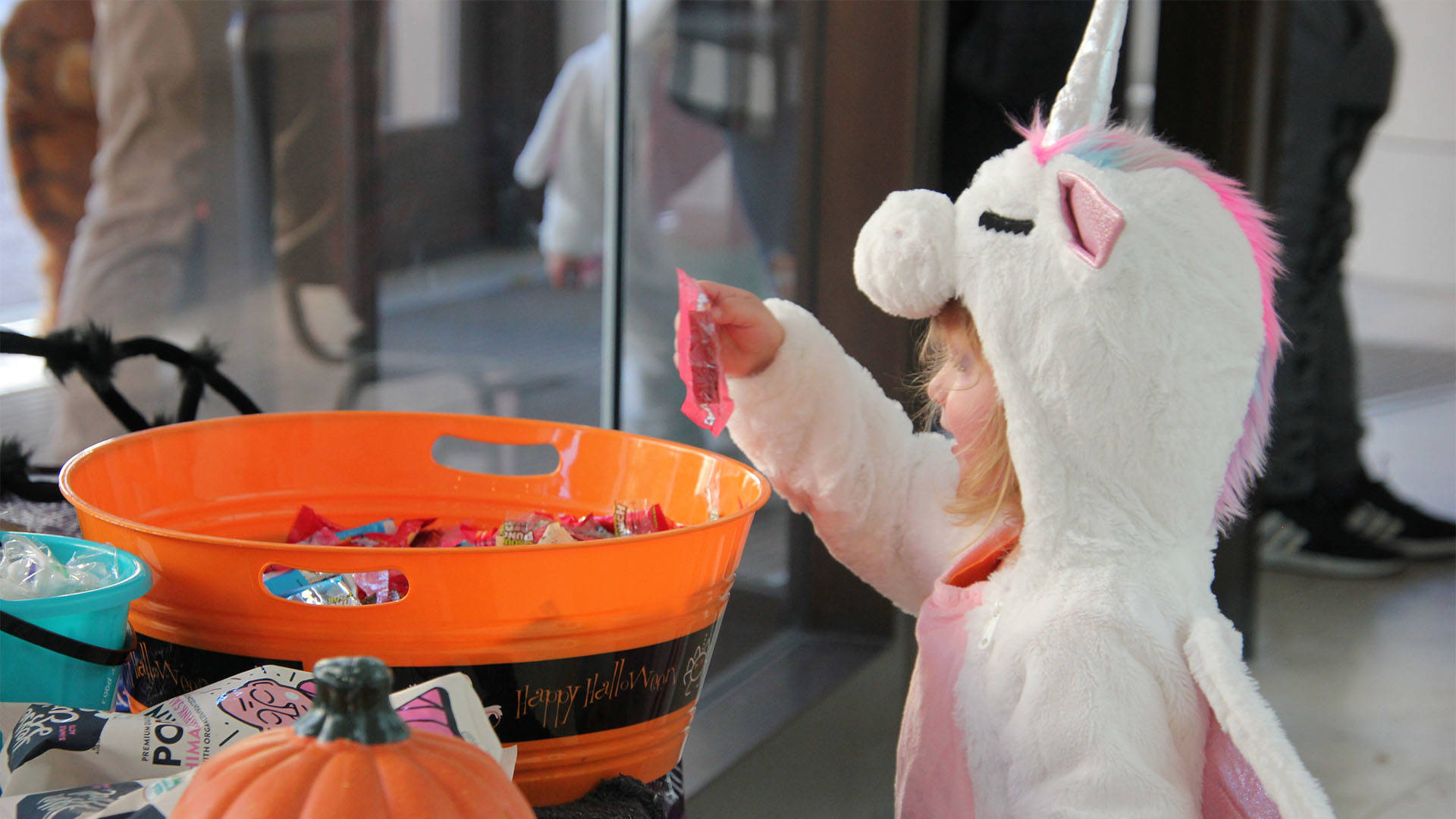 A young child in a white unicorn costume grabs a piece of candy from an orange bucket.