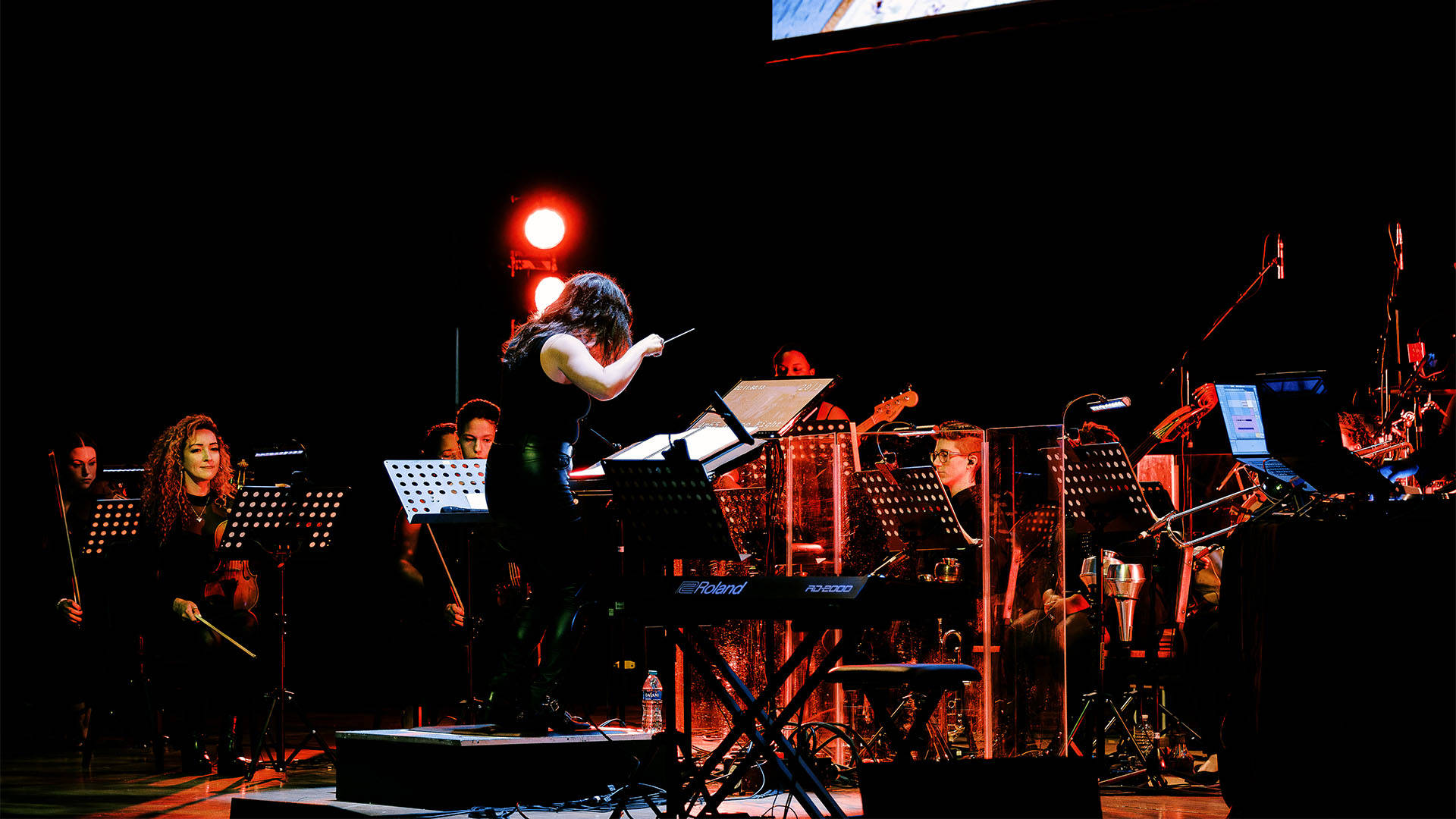 A woman with light complexion and long dark hair in a dark outfit conducting an orchestra.