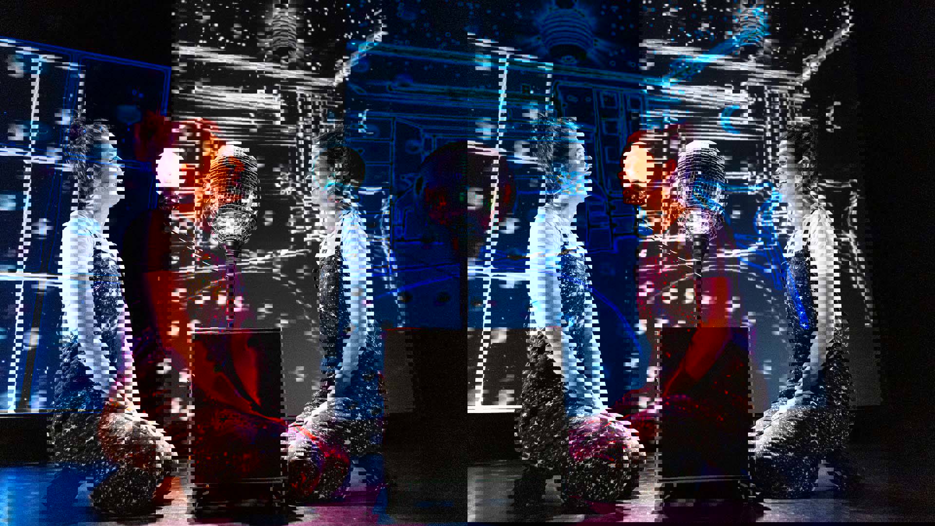 Two women in matching overalls with cosmic designs kneeling around a disco ball and glowing orb set to mimic a solar system model. The backgorund is dark with cyan digital patterns.