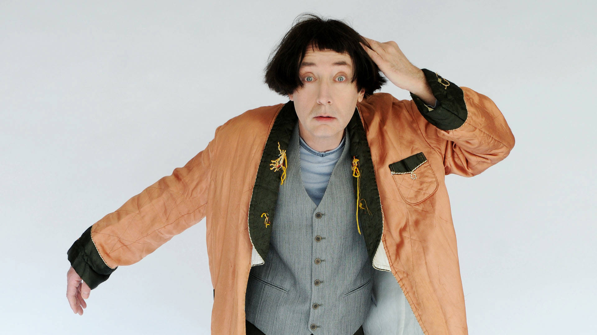 Emo Philips holds one hand out to his right and rests the other on his head. He has bright blue eyes and medium length straight black hair. He is wearing a bit orange robe and a vest