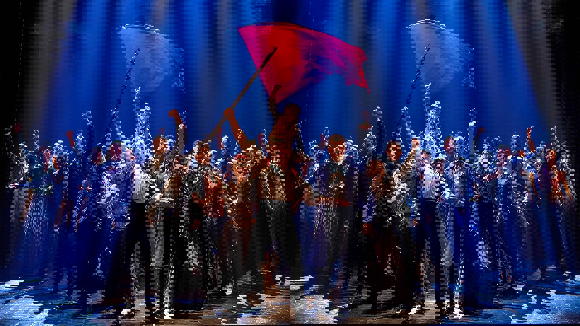 The cast of Les Miserables performing in a v formation and fists int he air. A red flag is waving above them.