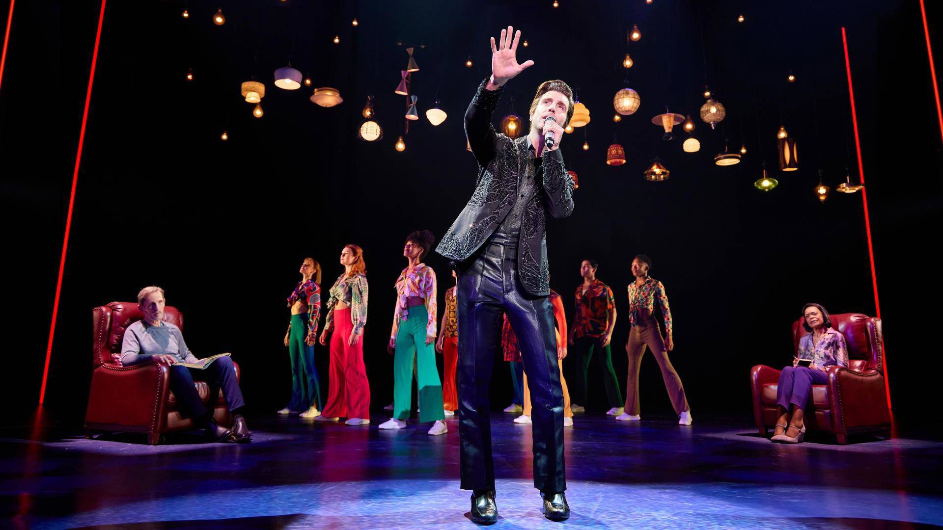 A man with light complexion, short brown hair, and a black outfit singing with a microphone. Performers in colorful clothing stand behind him. Two are sitting in plush red chairs.