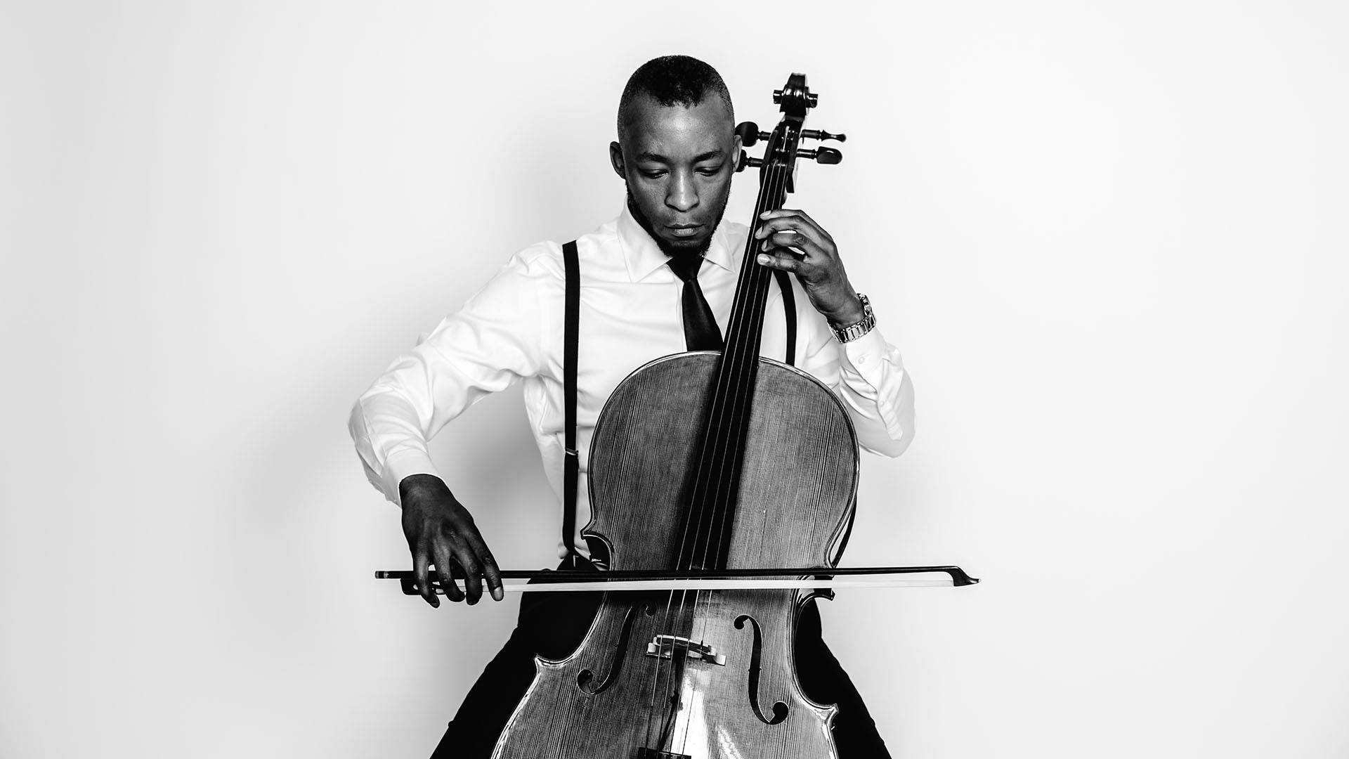 Headshot of Gabriel Royal in greyscale. He is a young handsome man wearing a white dress shirt, suspenders and a black tie. He is seated and playing a cello