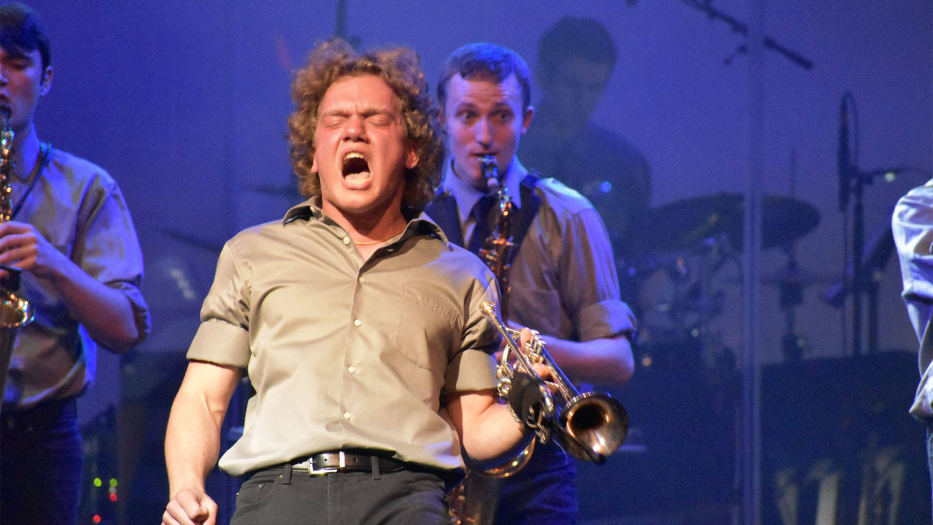 A high school boy in a beige shirt in performance holding a trumpet and yelling with his eyes closed