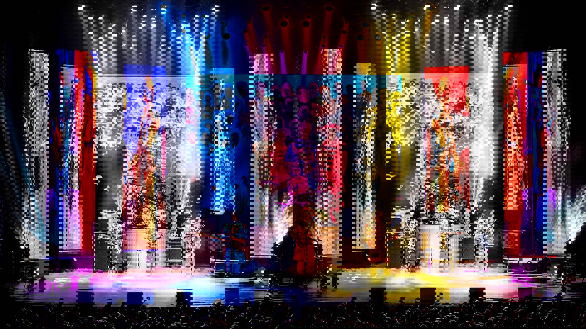 Four Beatles impersonators performingon a colorful stage. They are wearing Seargent Peppers outfits with the album cover projection behind them.