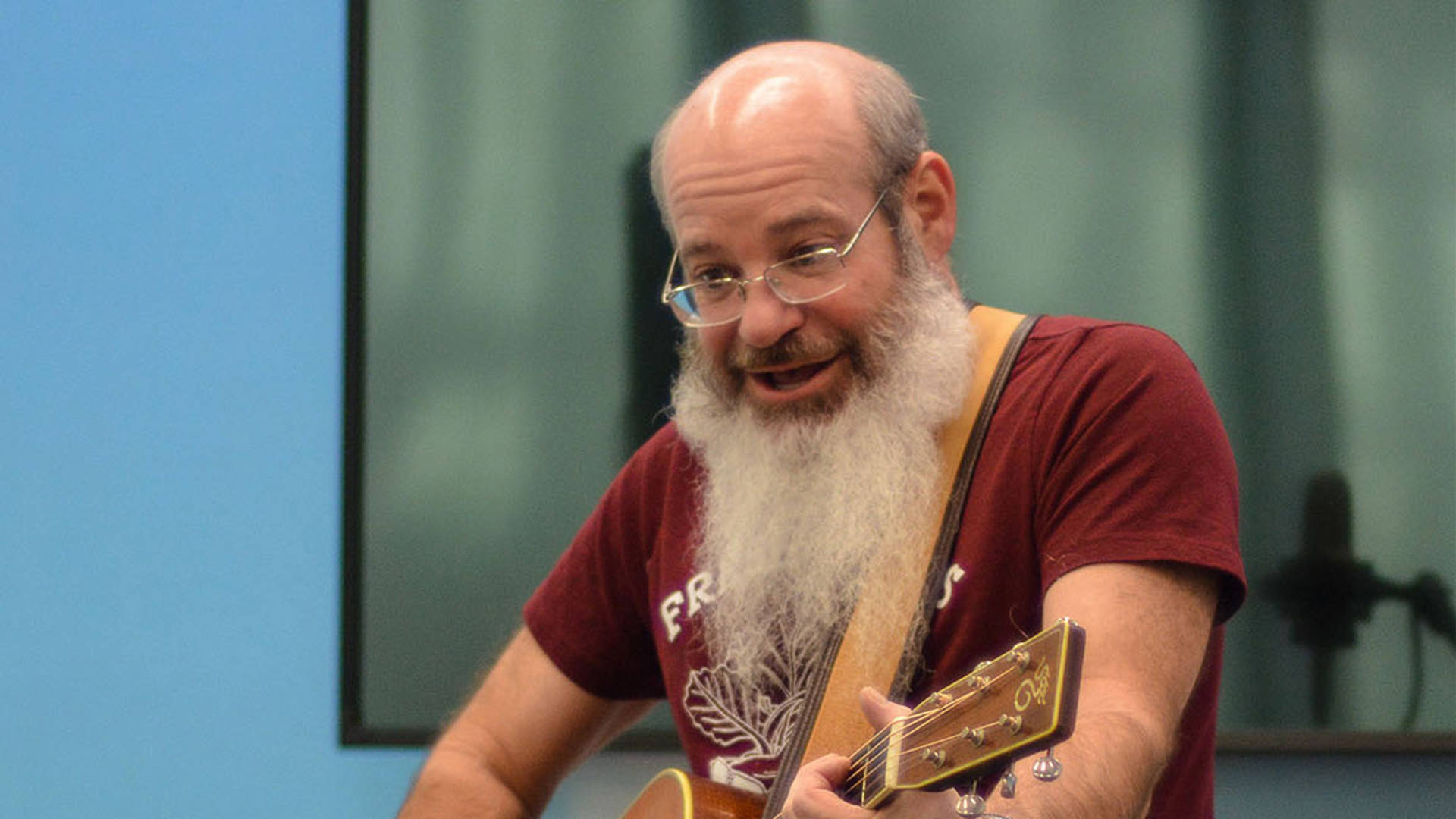 David Laundau is holding a guitar and singing. He is a caucasian man with a bald head, grey hair, a long beard, glasses and a red shirt on.