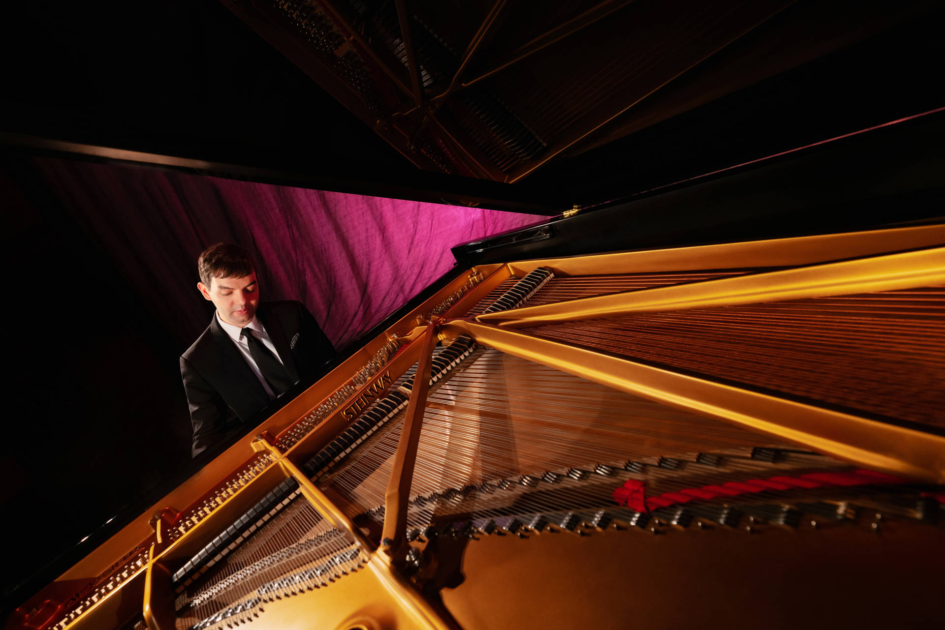 A man with light complexion and brown hair plays an open grand piano in front of a pink stage curtain.