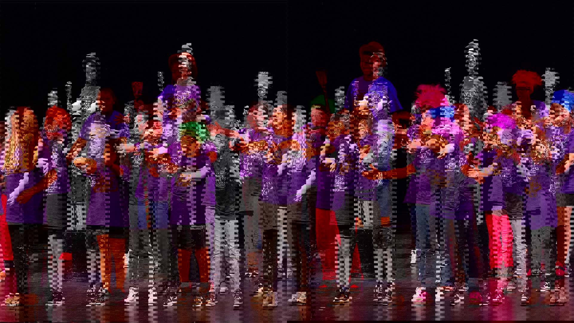 Young students in purple shirts and colorful headwear performing on a dark stage. 