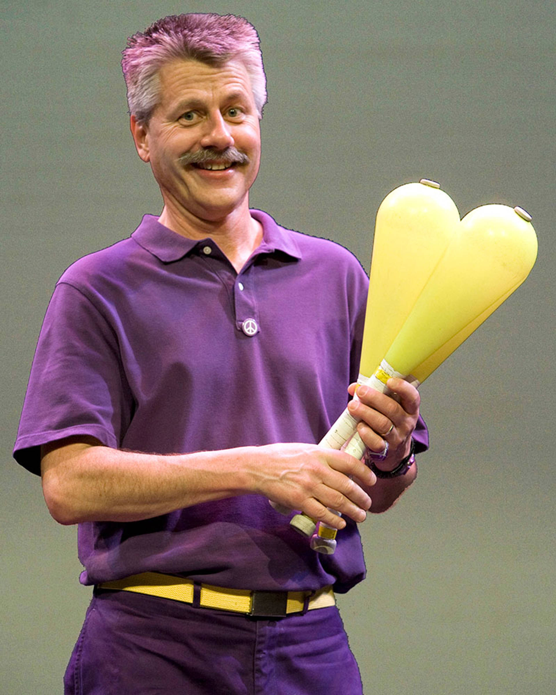 A man dressed in all purple with short grey hair and a moustache holding yellow juggling pins