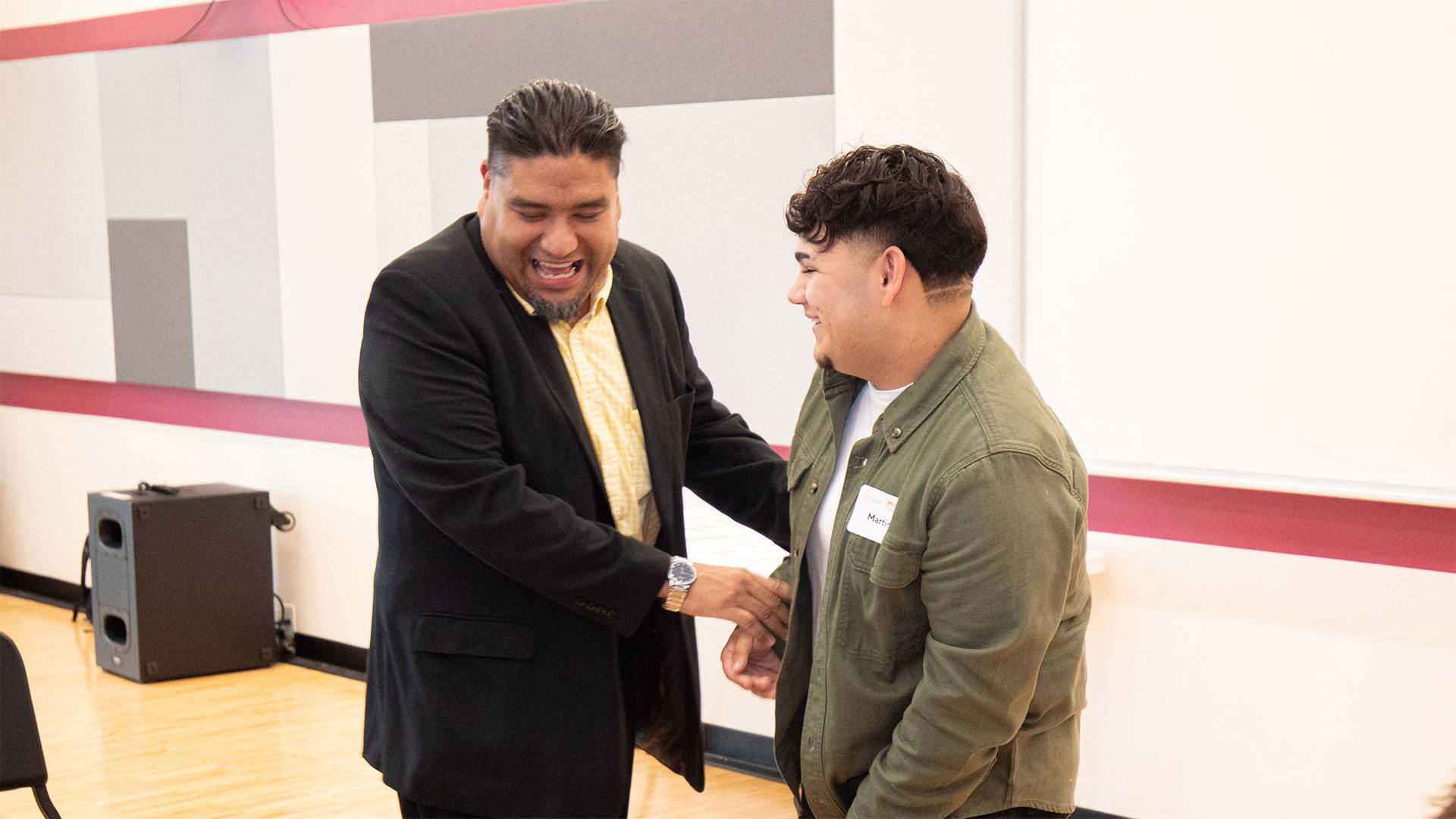 Two men laughing together. The man on the left has medium complexion and is older than the man on the right. He is wearing a suit jacket. The younger man on the right has medium complexion, brown hair and green jacket.