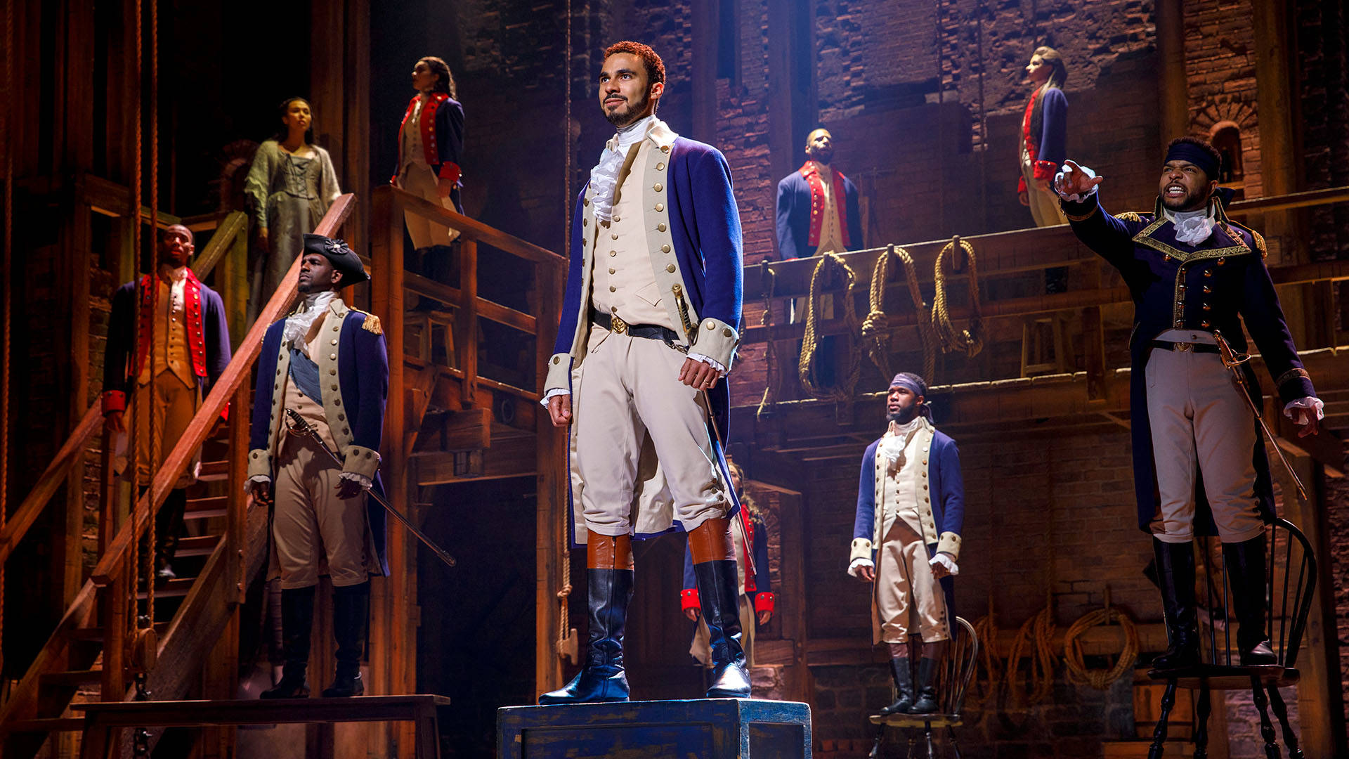Cast members of Hamilton stand tables, chairs and stairs. The are dressed in revolutionary war outfits.