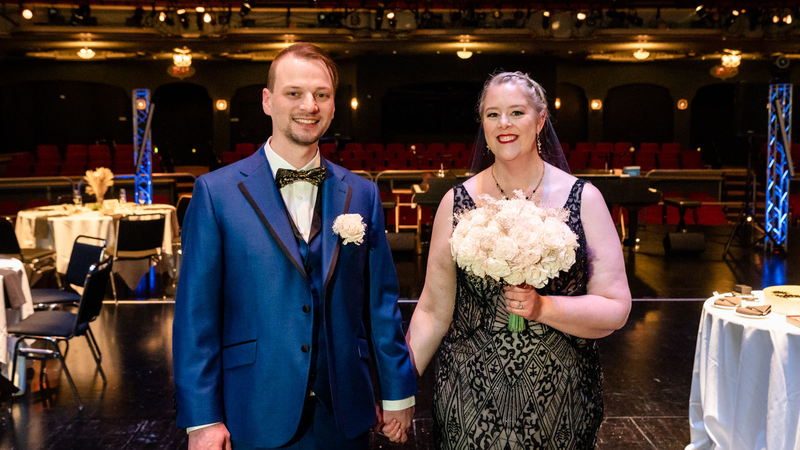 A man in a blue suit and a woman in a beige and black dress holding hands and smiling for a photo. The woman is holding a bouquet of white roses.