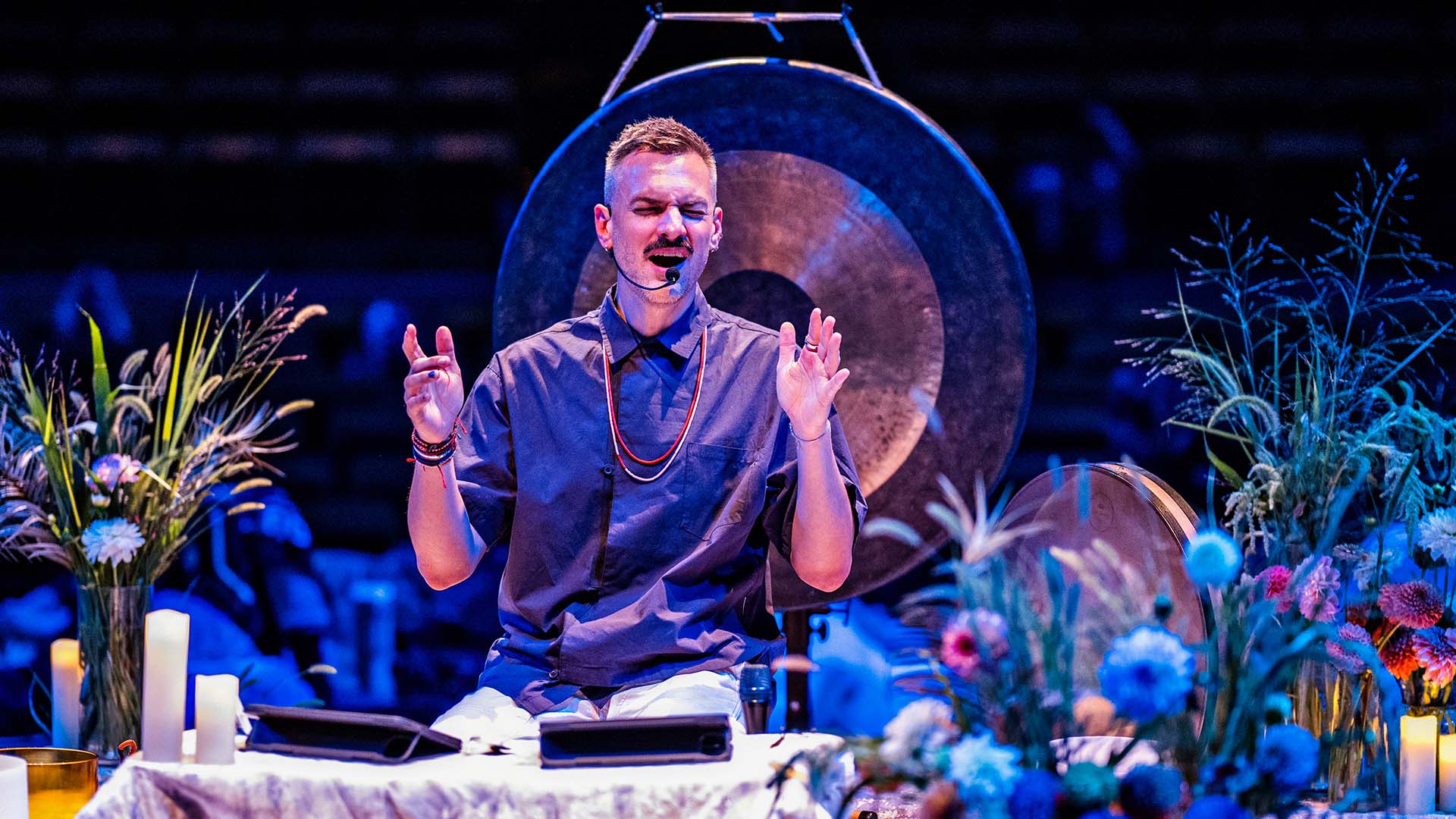A white man with short blonde hair in a black button up shirt sits on a small stage surrounded by vases of flowers. A large gong is behind him with candles surrounding him. Both of his hands are raised as he sings into a headset microphone.