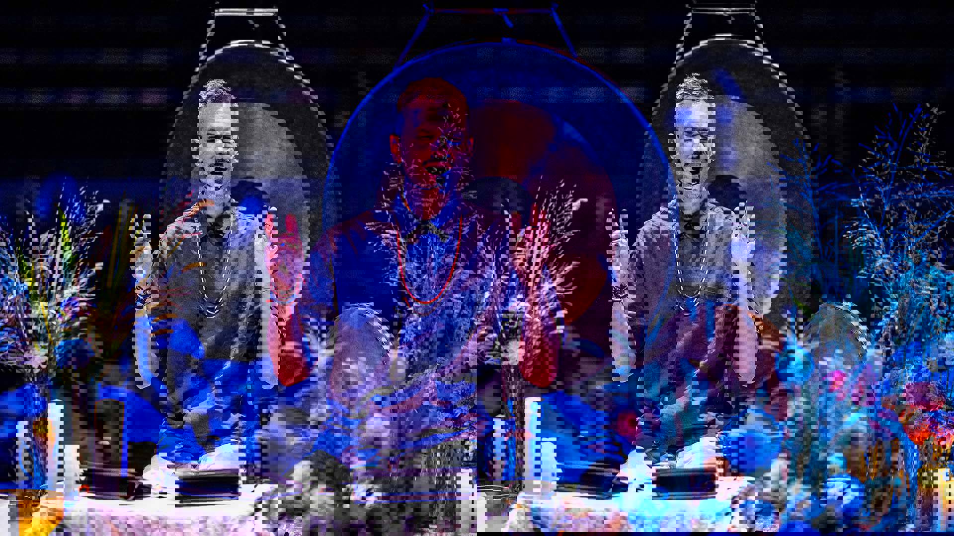 A white man with short blonde hair in a black button up shirt sits on a small stage surrounded by vases of flowers. A large gong is behind him with candles surrounding him. Both of his hands are raised as he sings into a headset microphone.