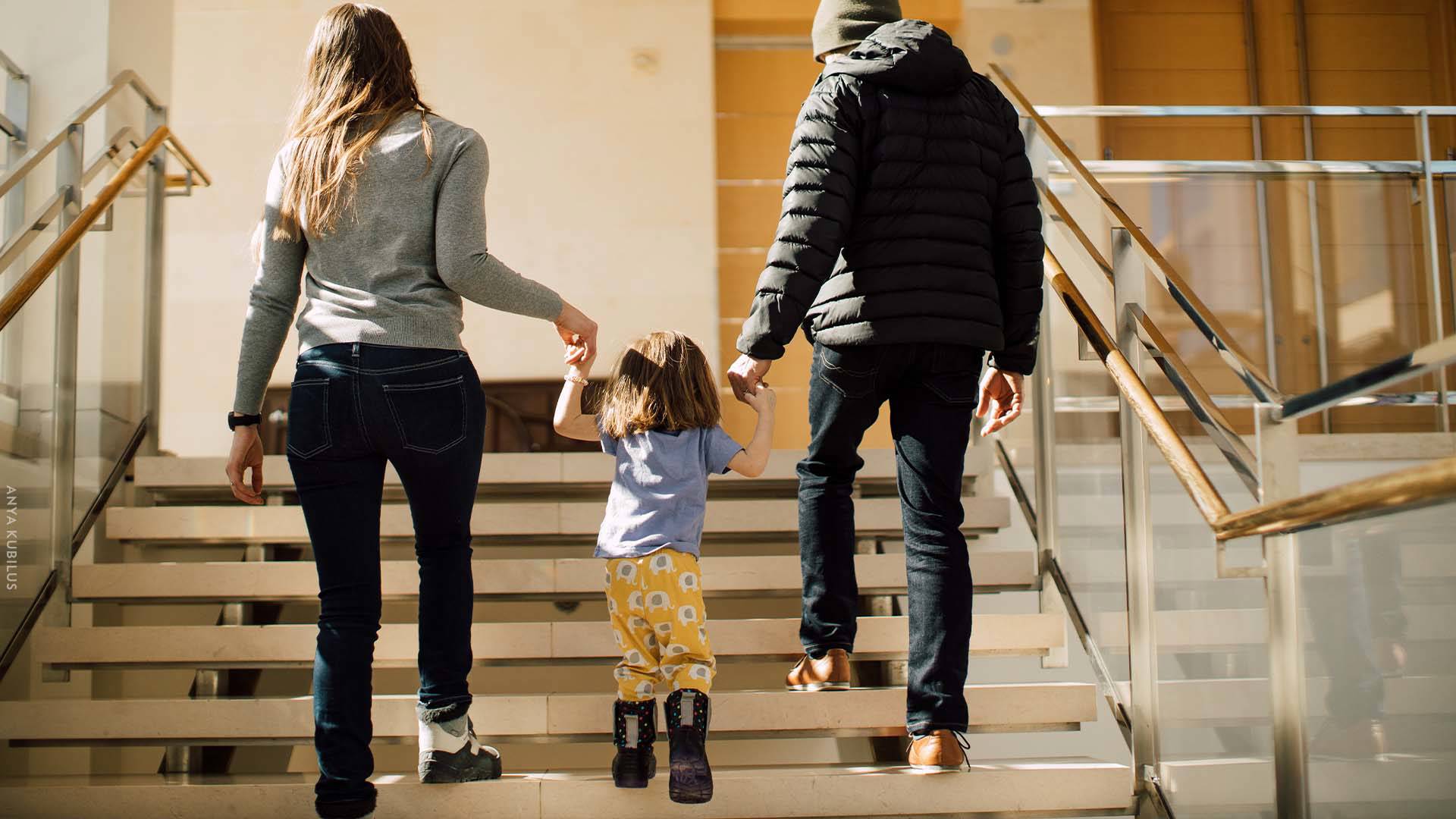Two parents holding their child's hands as they walk up stairs in Overture Hall Main Lobby.